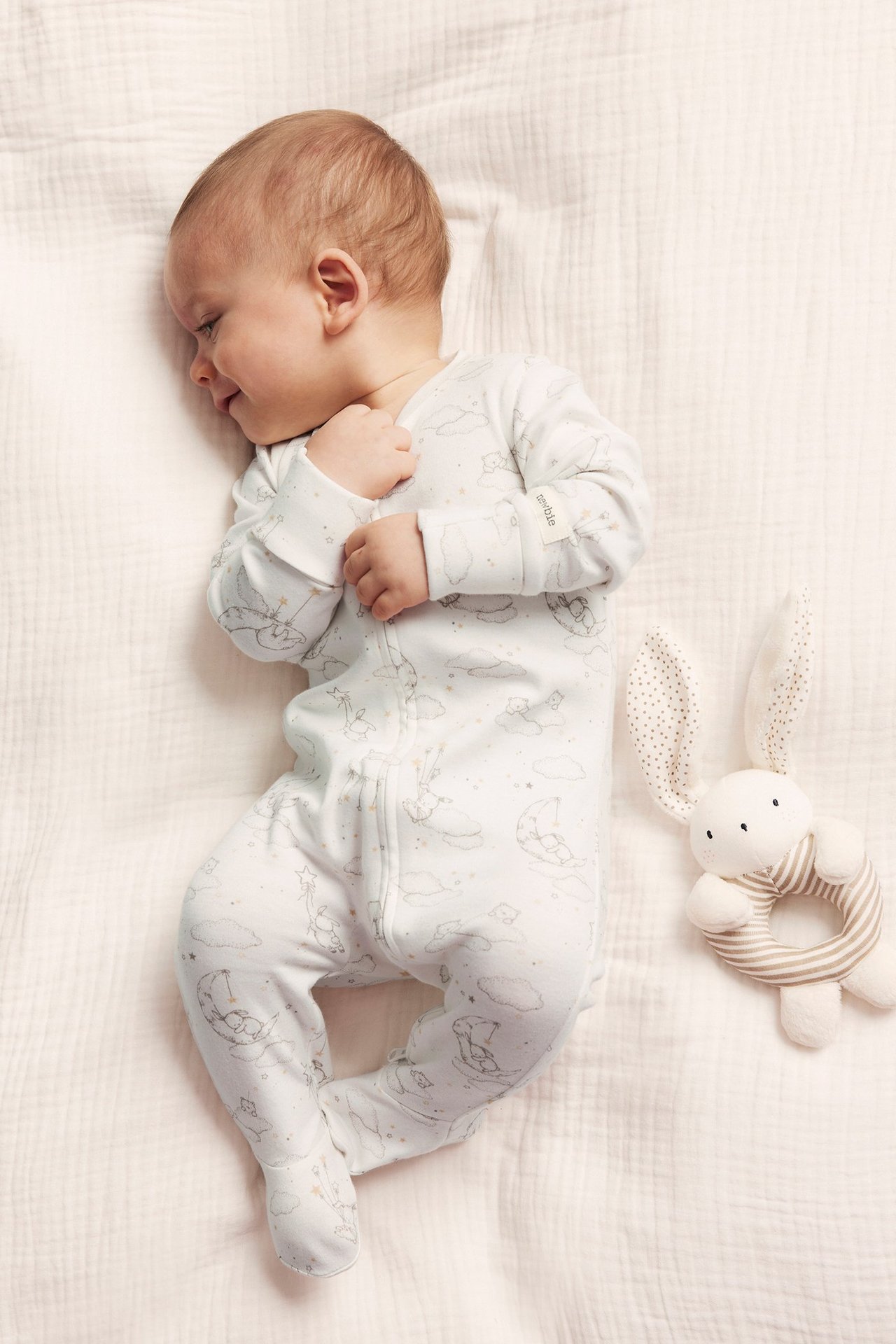 Top view of a baby in an off-white cotton sleepsuit with bear and moon pattern, zipper. Bunny rattle beside.