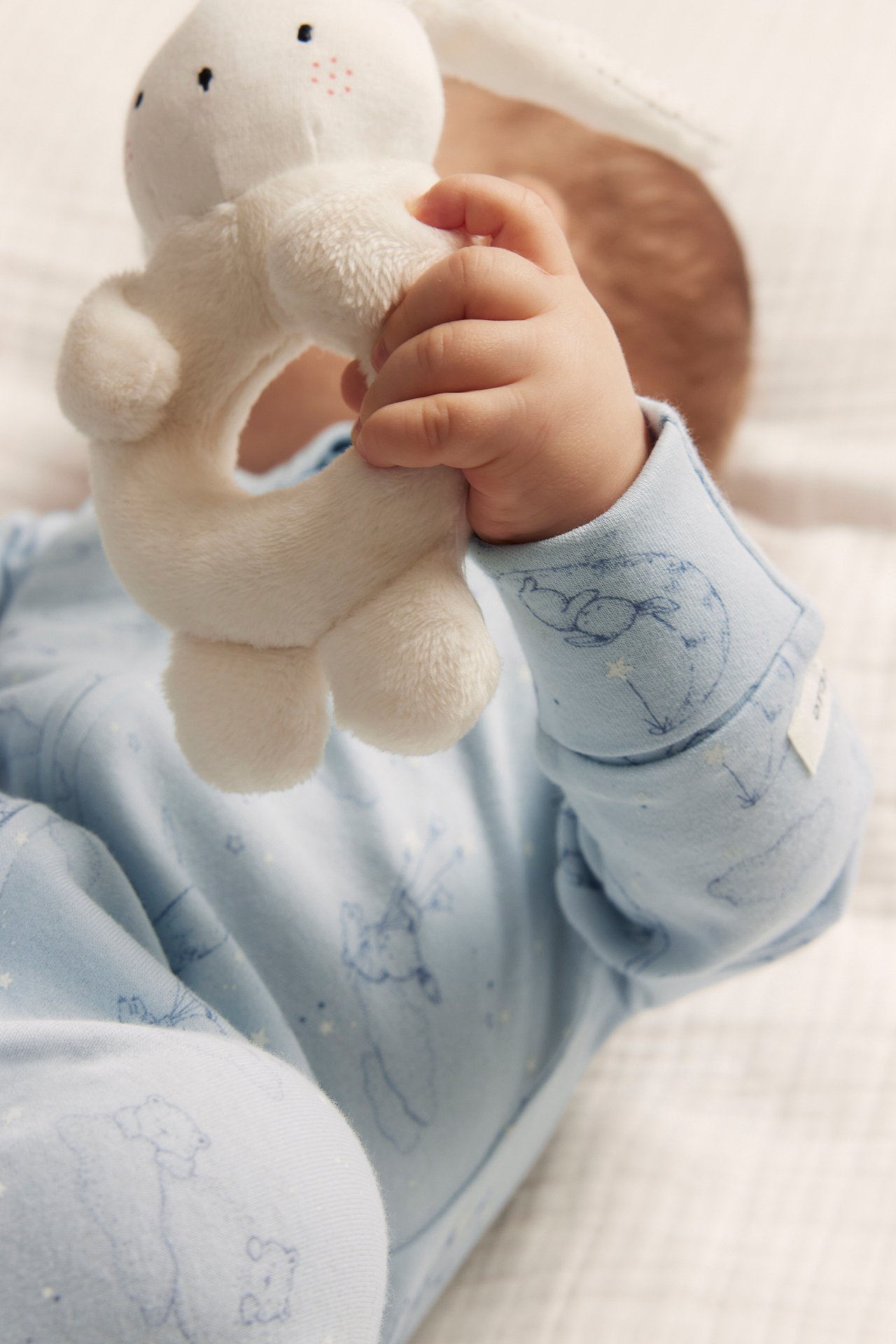 Close-up of a baby in a light blue patterned cotton pyjama, holding a white plush bunny rattle.