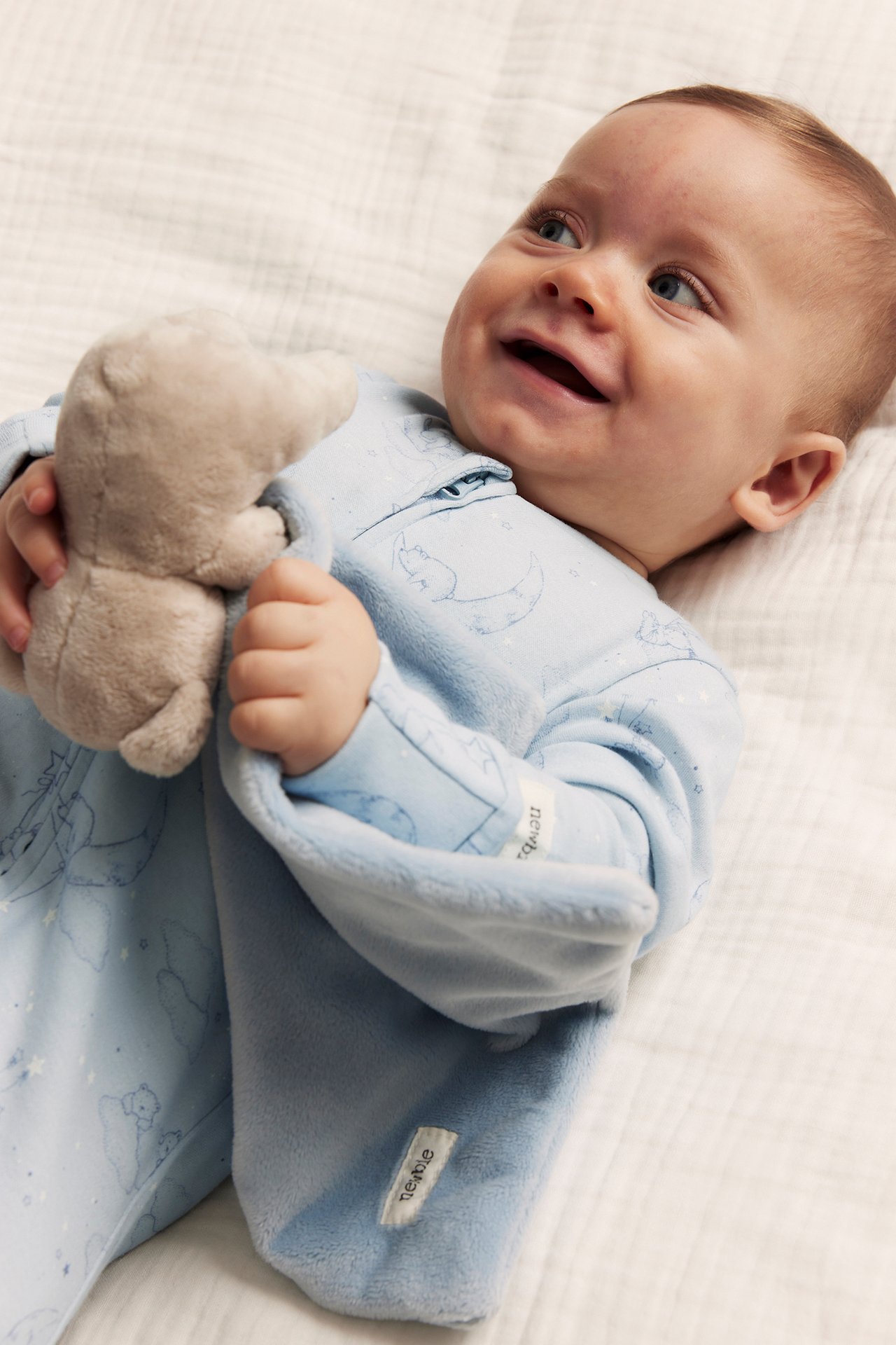 Close-up of a smiling baby wearing light blue patterned cotton pajamas with a zipper, holding a small stuffed animal.