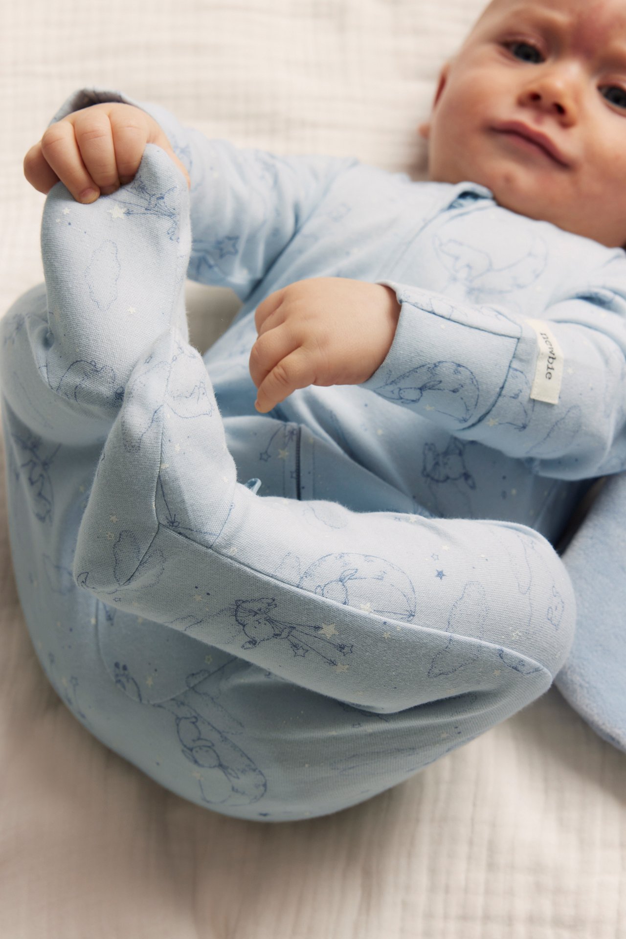Close-up of baby in light blue cotton pajamas with bear, rabbit, star, moon pattern, holding foot.
