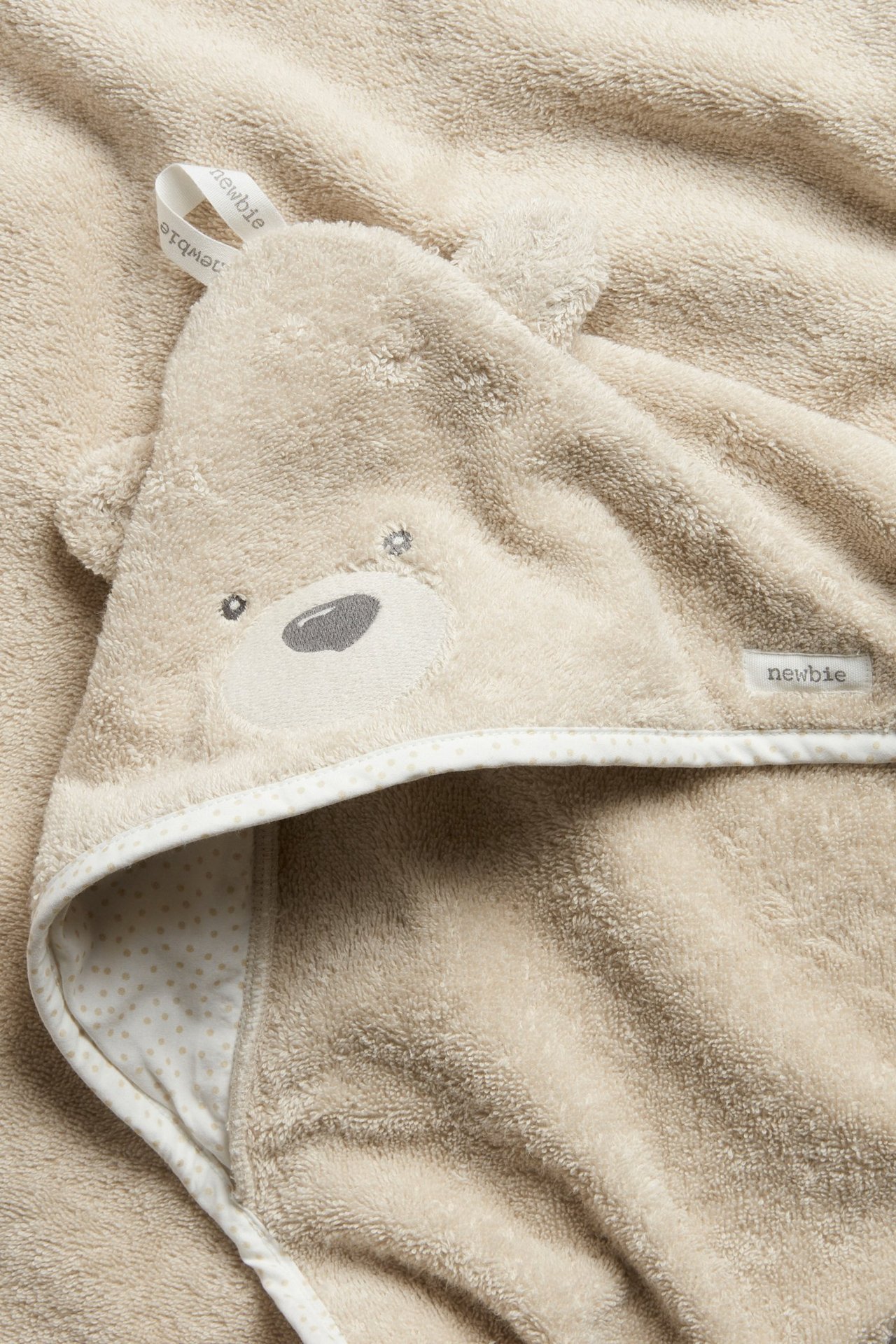 Close-up of a beige terry baby hooded bath towel with bear ears, embroidered details, and a dotted edge.