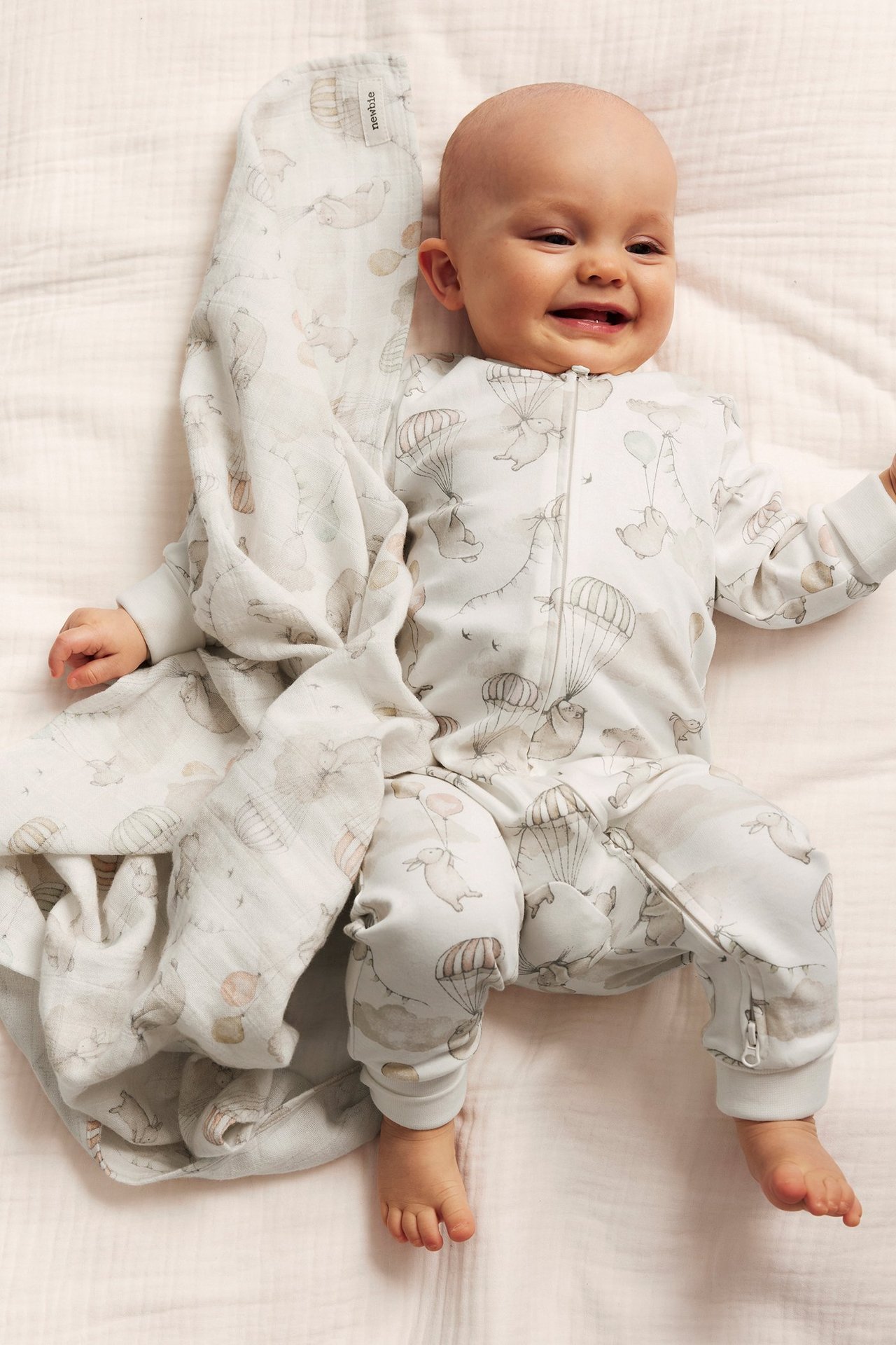 Smiling baby in an offwhite patterned romper with bunnies, parachutes, and a two-way zip. Matching blanket beside.