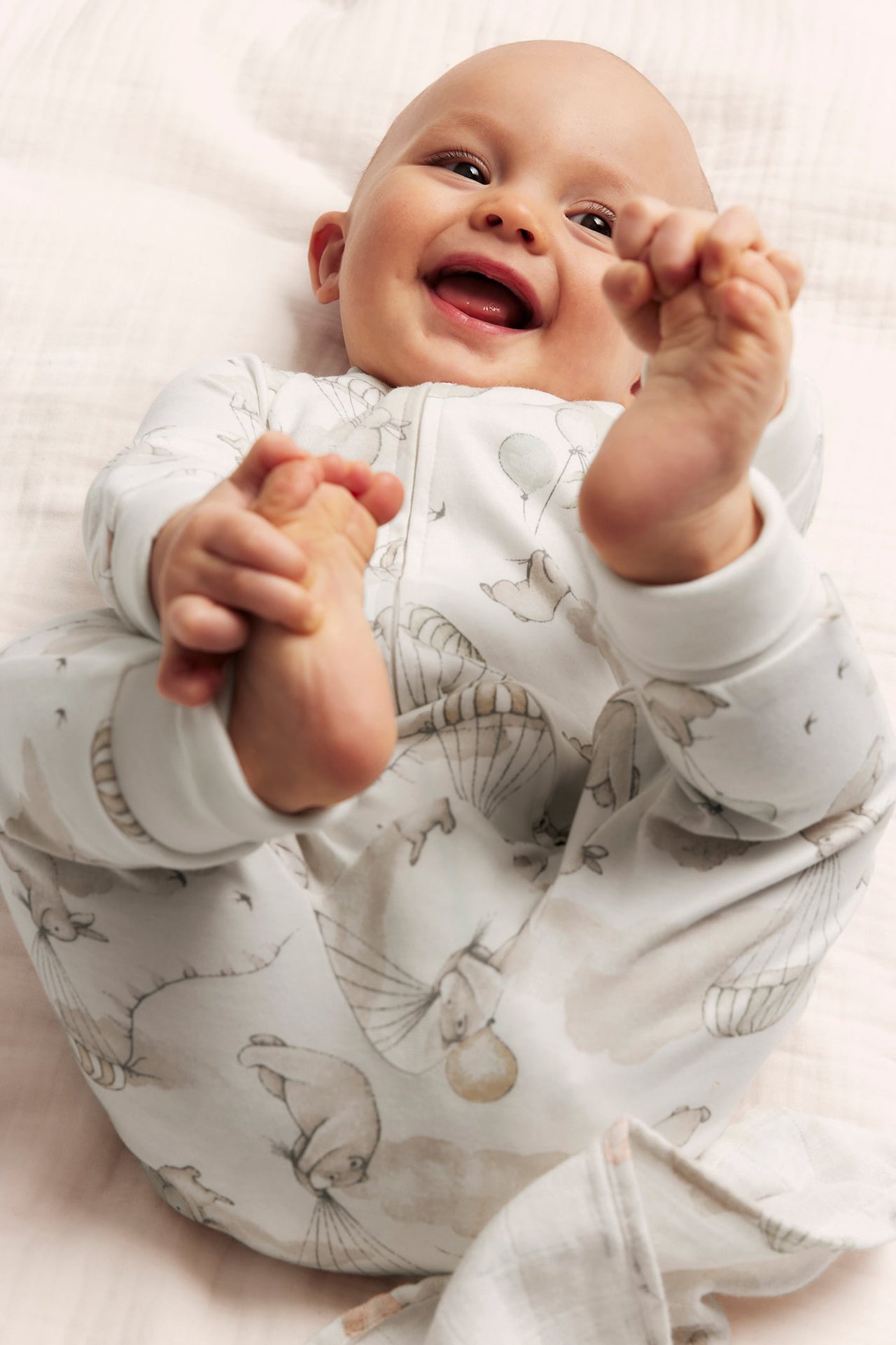 Nahaufnahme eines glücklichen Babys in einem cremeweißen gemusterten Baumwoll-Schlafanzug mit Zwei-Wege-Reißverschluss, das seine Füße hält.