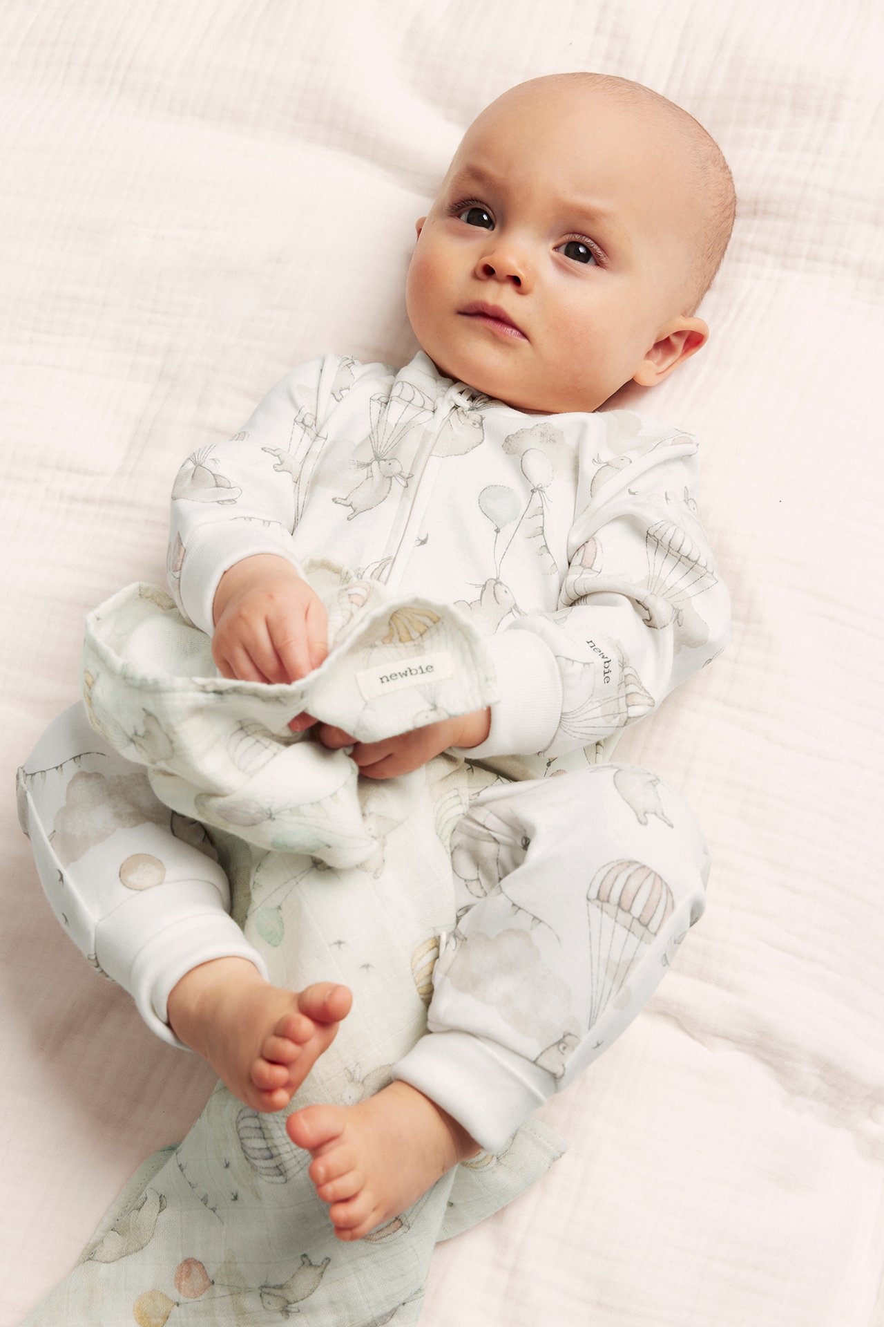 Vorderansicht eines Babys in einem cremeweißen, tiermusterbedruckten Baumwoll-Strampler mit Reißverschluss, hält ein Tuch.