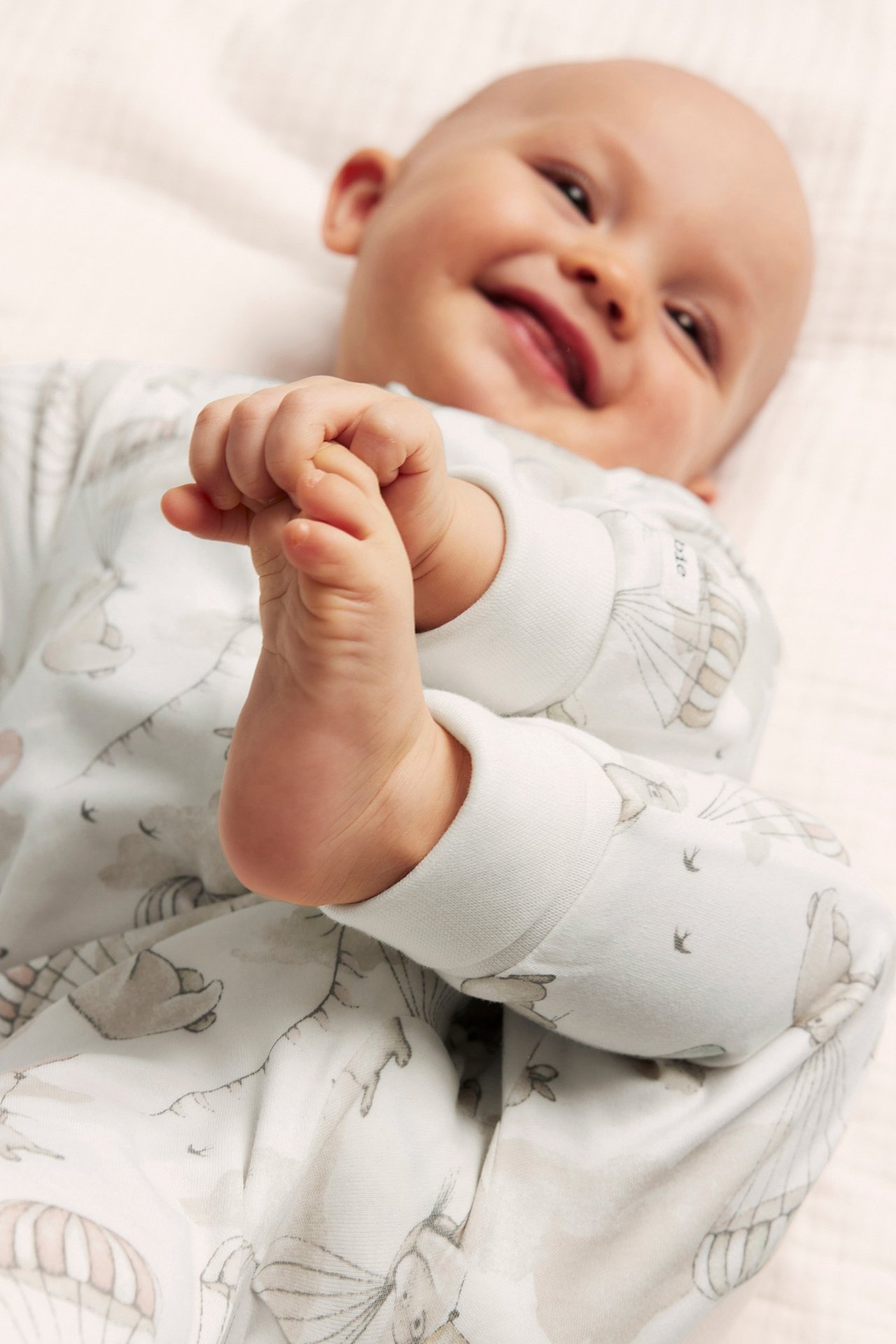 Close-up of a smiling baby in an off-white cotton romper with grey animal and hot air balloon pattern.