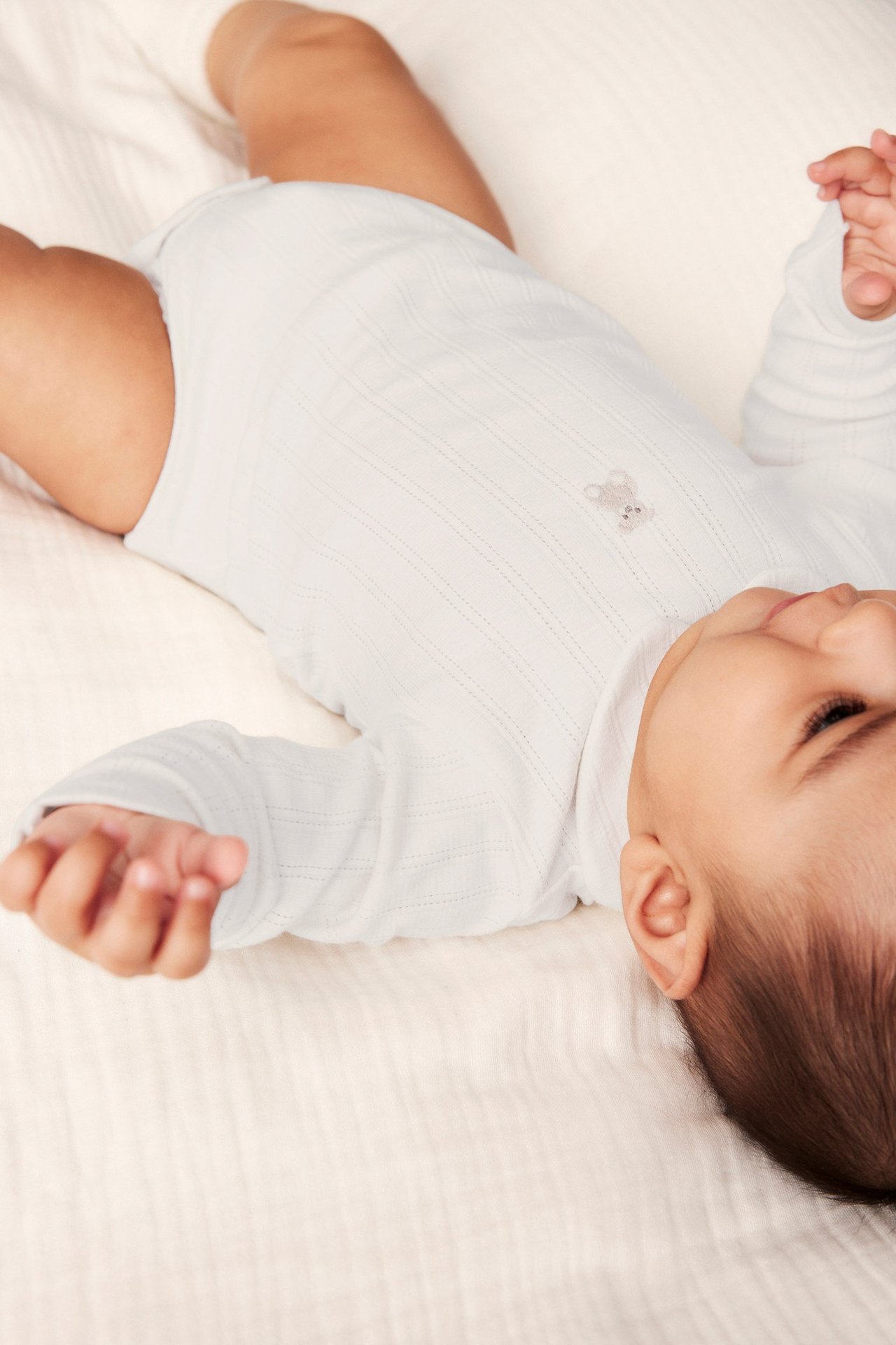 Front view of a baby wearing an offwhite pointelle rib long-sleeved bodysuit with teddy bear embroidery.