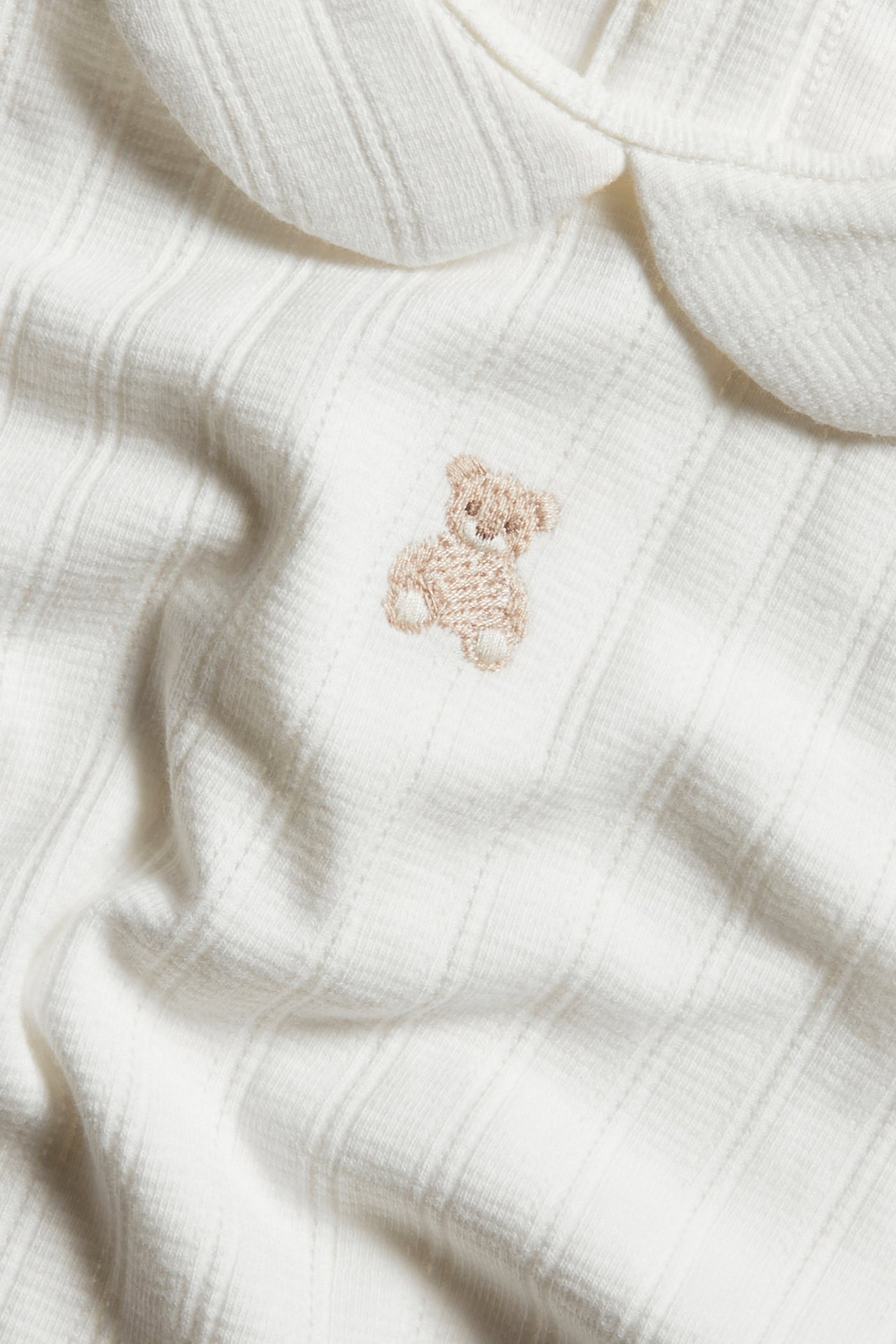Nahaufnahme eines cremeweißen gerippten Babybodys mit Kragen und Teddybär-Stickerei.