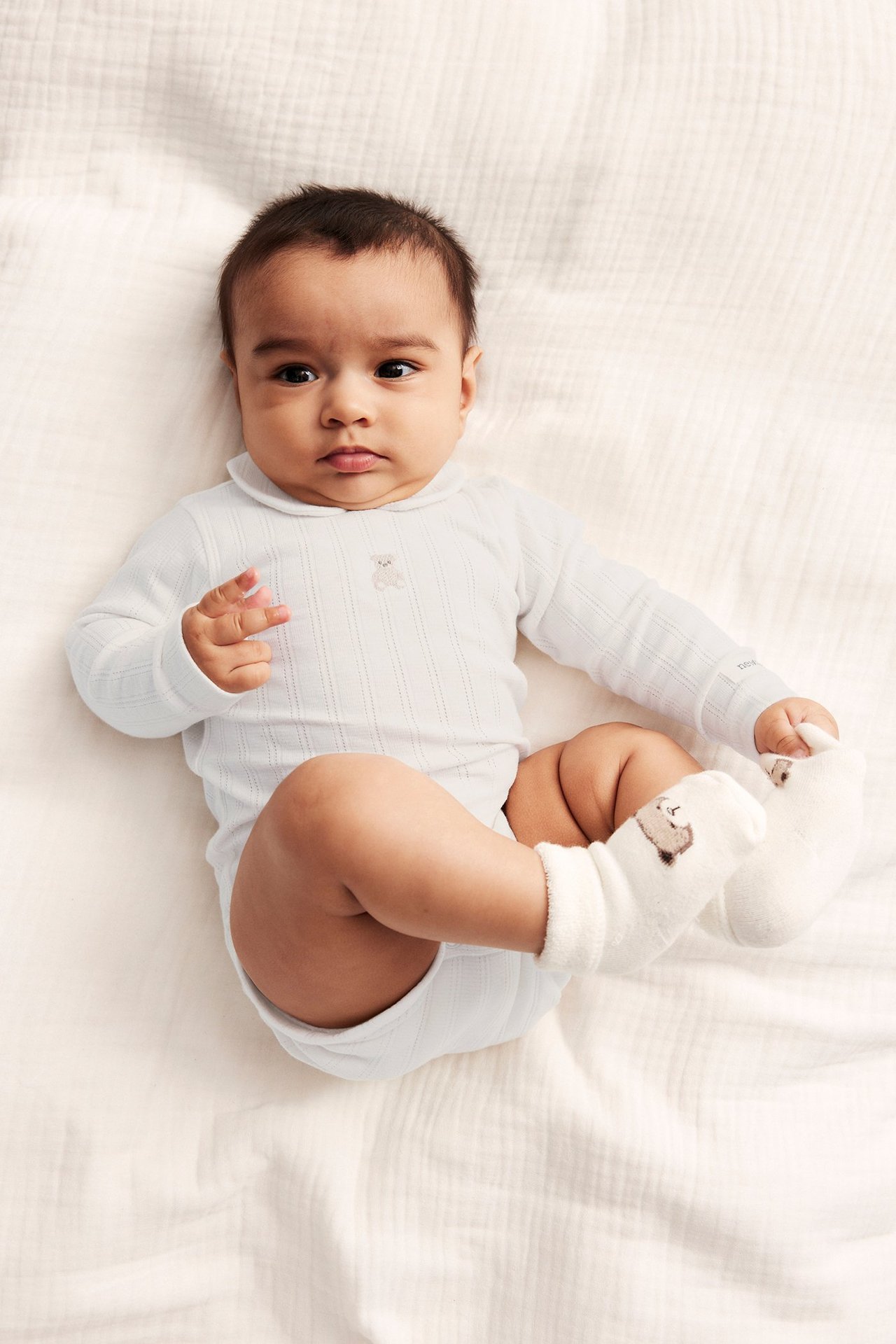 Top-down view of a baby in an off-white pointelle bodysuit with collar, teddy embroidery, and teddy socks.