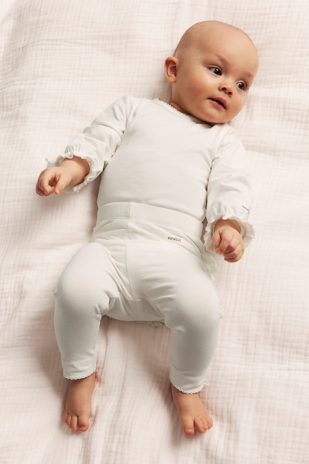 Top-down view of a baby wearing an offwhite long-sleeved bodysuit with ruffled cuffs and matching trousers.