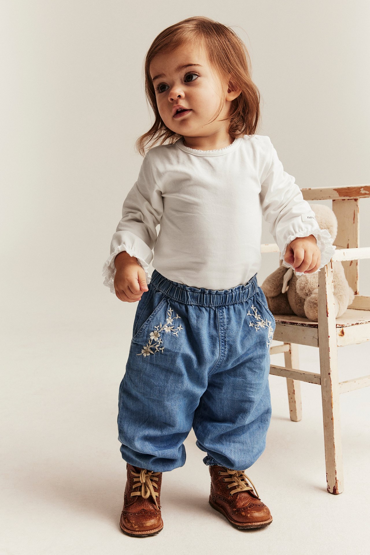 Vorderansicht eines Babys in einem cremeweißen Langarmbody mit Rüschenmanschetten, blauen bestickten Hosen und braunen Schnürstiefeln.