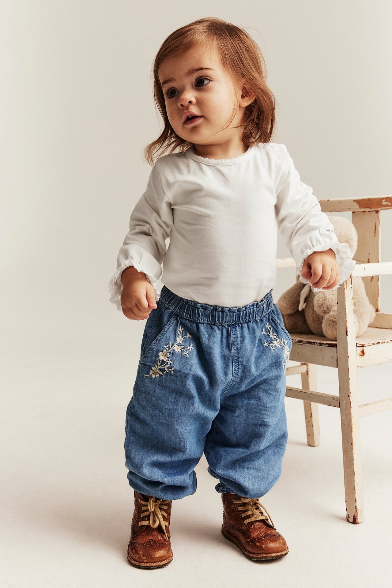 Front view of a baby in an offwhite ruffled long-sleeved bodysuit, blue embroidered pants, and brown lace-up boots.