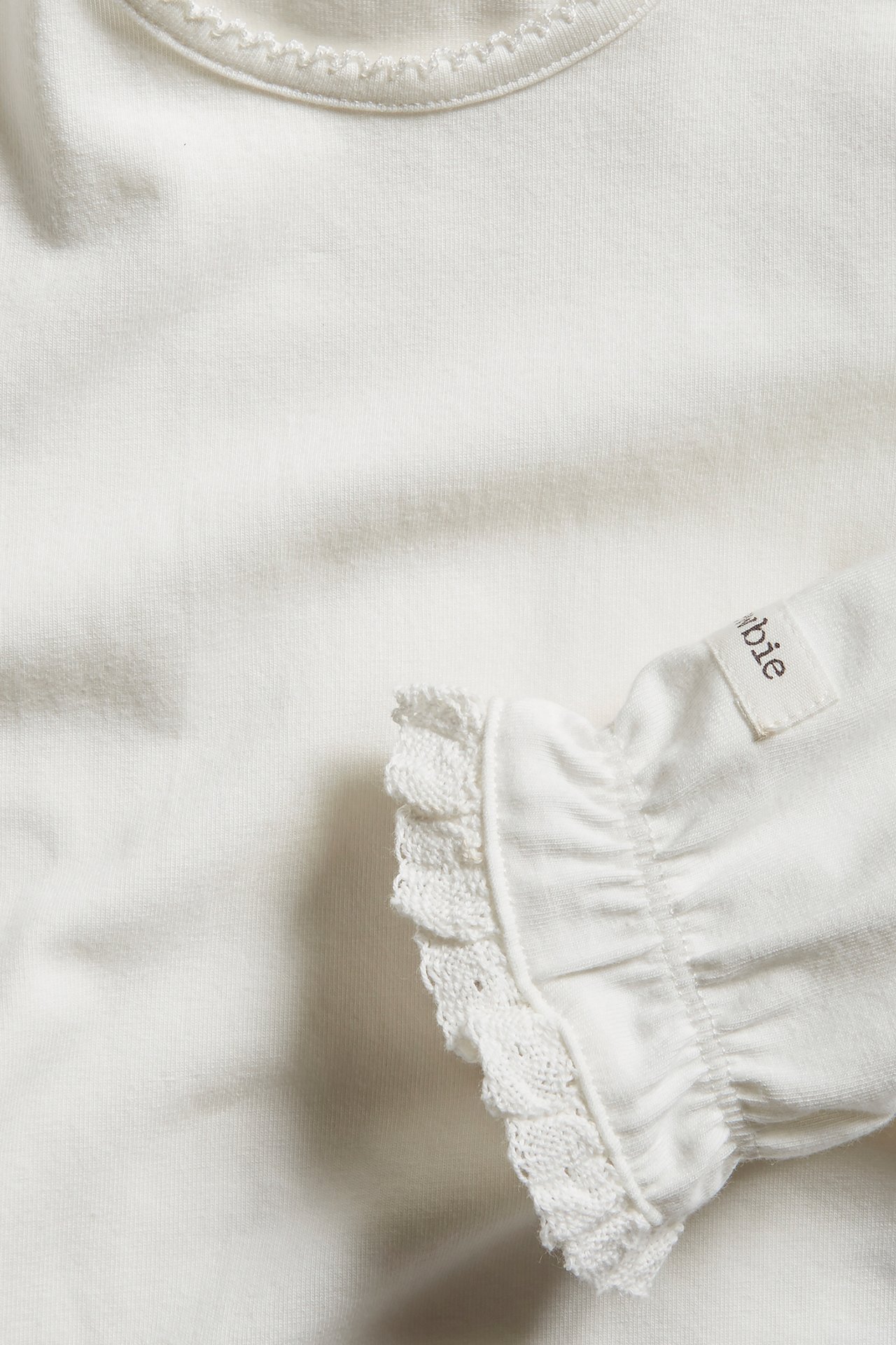 Nahaufnahme eines cremefarbenen Baby-Bodys mit Picot-Ausschnitt und Rüschenärmelbündchen mit Spitze.