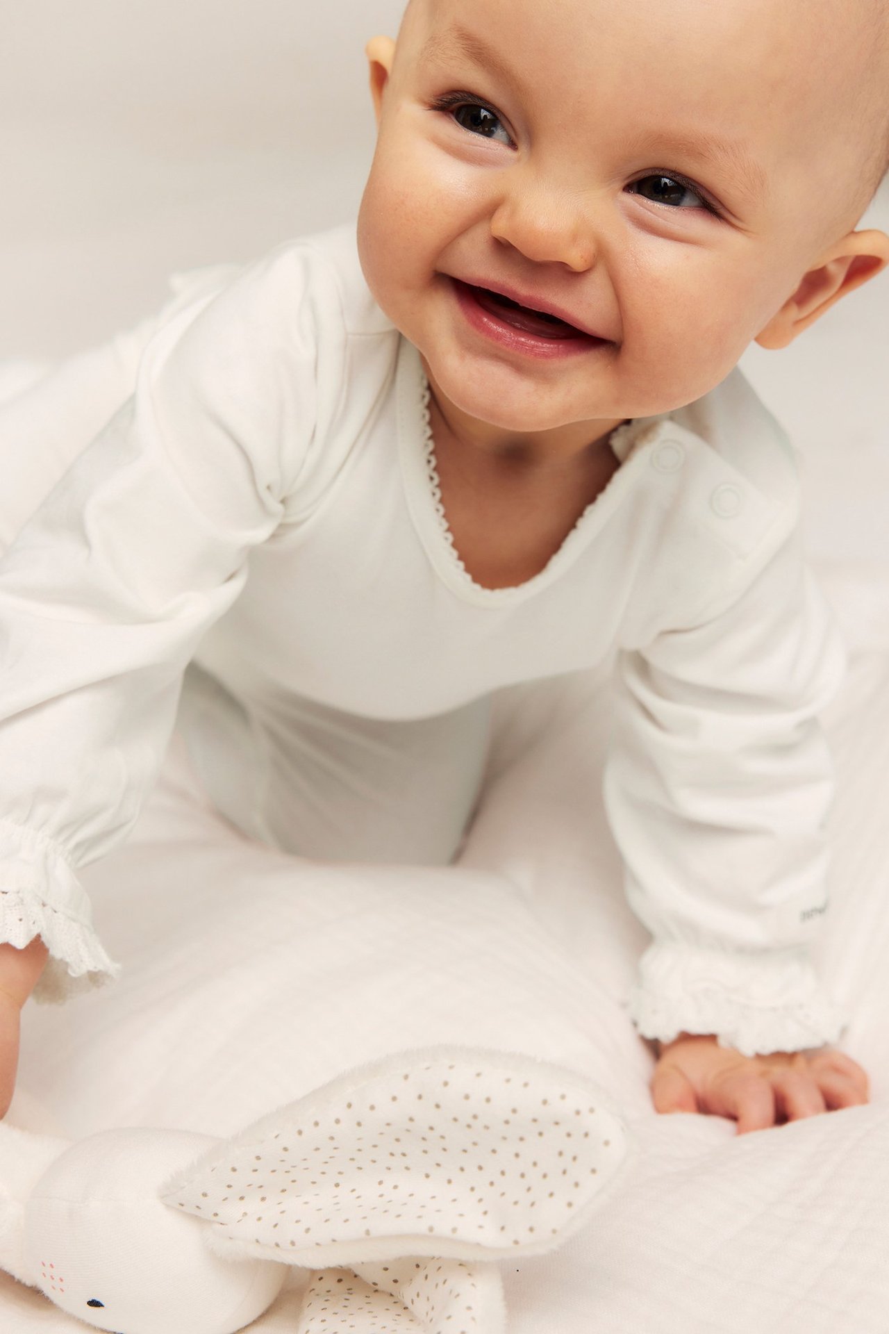Smiling baby in an off-white long-sleeved bodysuit with picot trim, lace cuffs, and a white bunny toy.