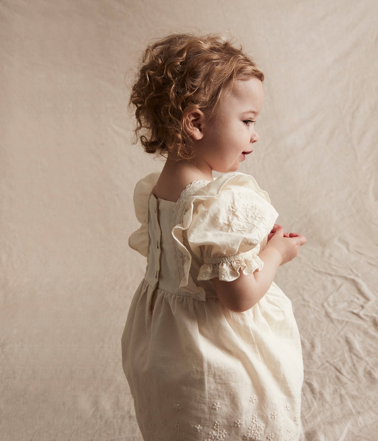Side view of a baby wearing an off-white embroidered cotton dress with puff sleeves and back buttons.