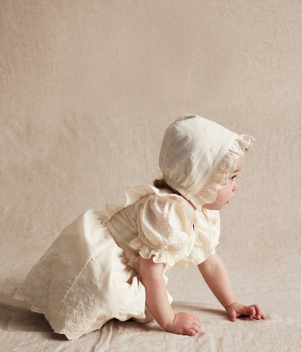 Side view of a baby crawling in an offwhite embroidered cotton dress with puff sleeves and matching bonnet.