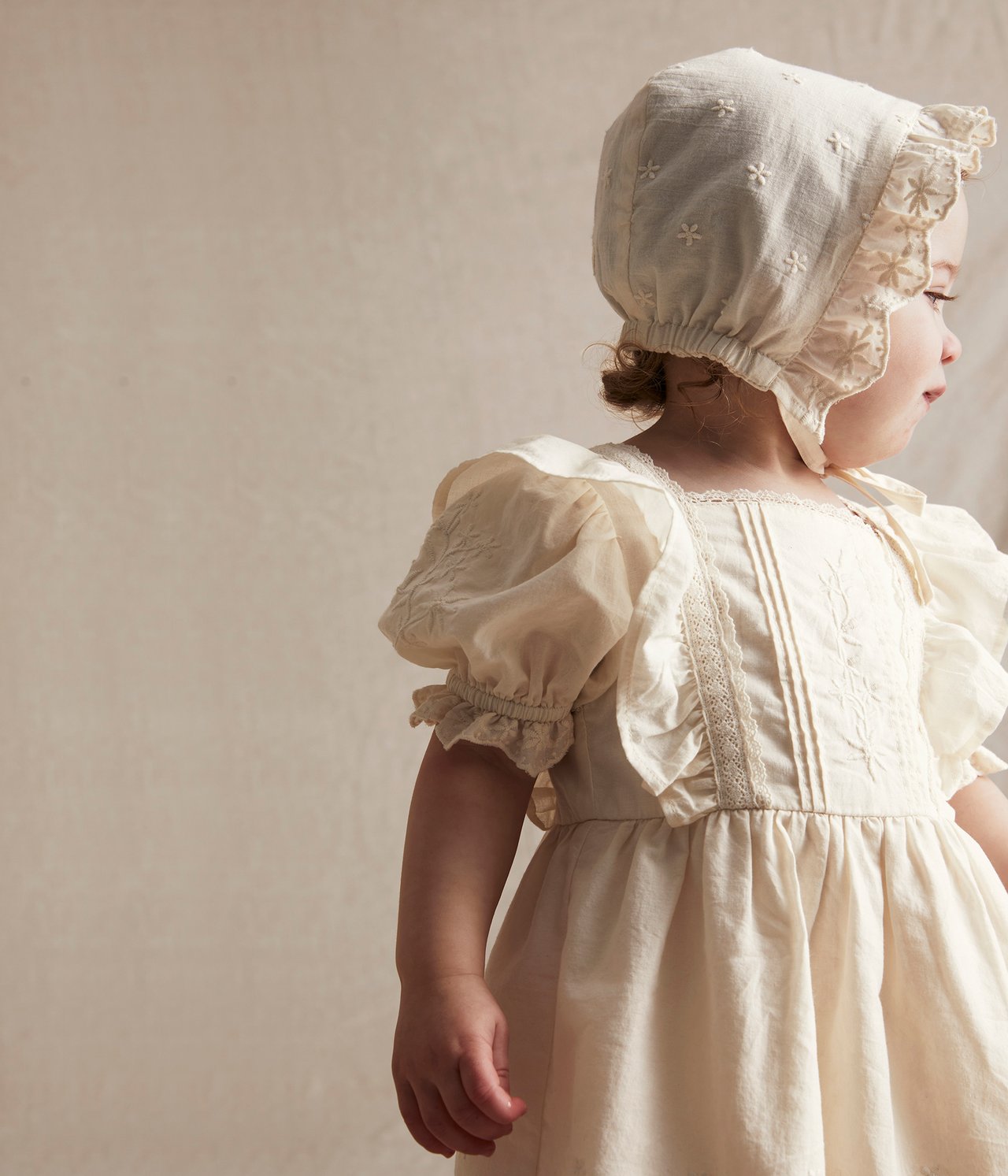 Side view of a baby wearing an off-white embroidered cotton dress with puff sleeves and a matching bonnet.