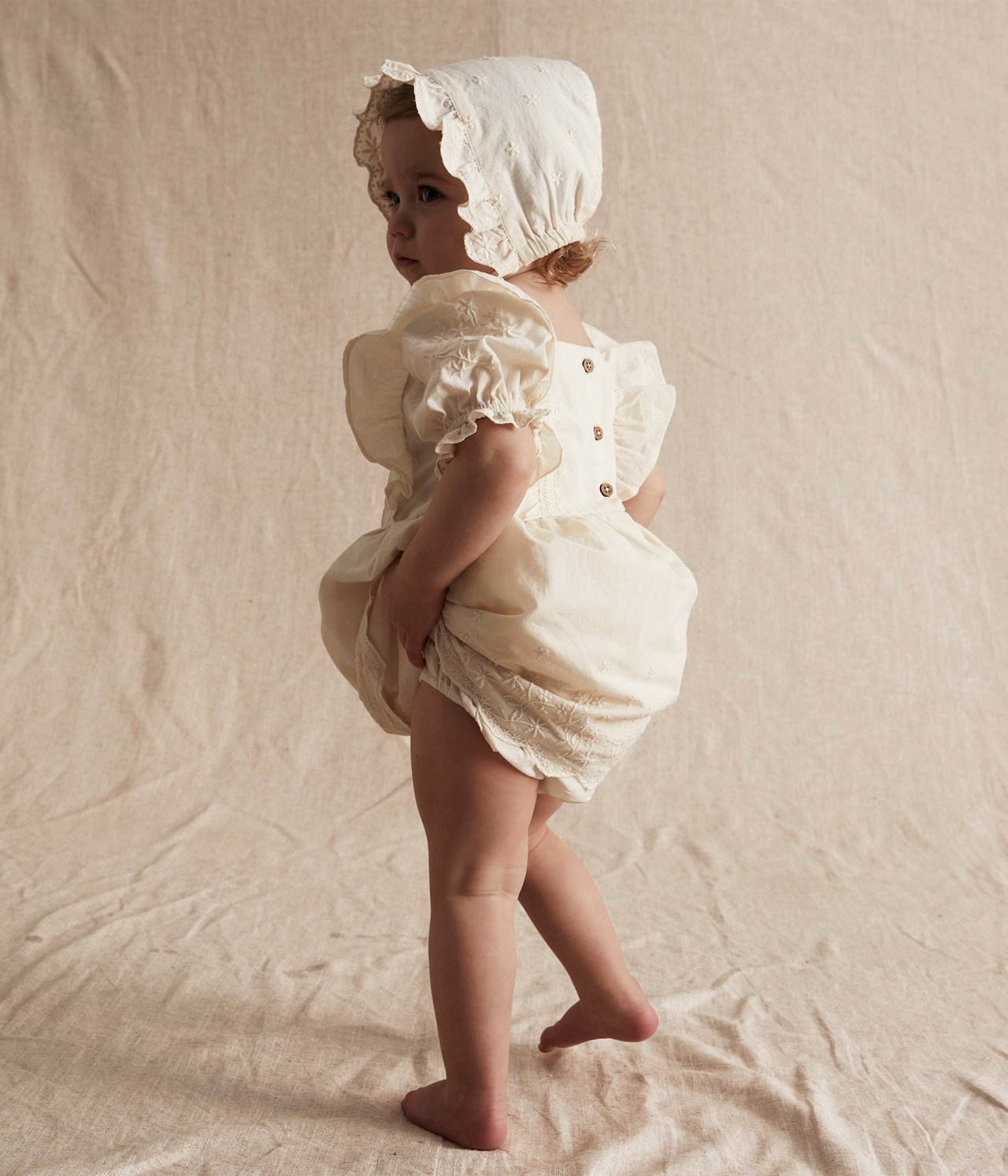 Side view of a baby in an off-white embroidered cotton dress, puff shorts, and lace-trimmed bonnet.