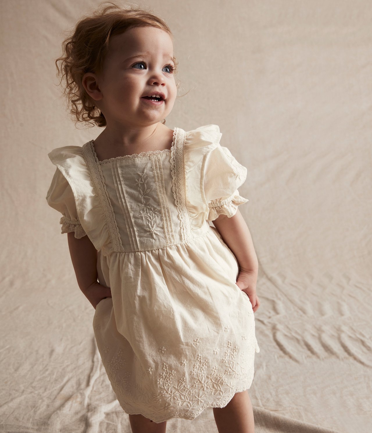 Front view of a baby wearing an off-white embroidered cotton dress with lace details and puff sleeves.