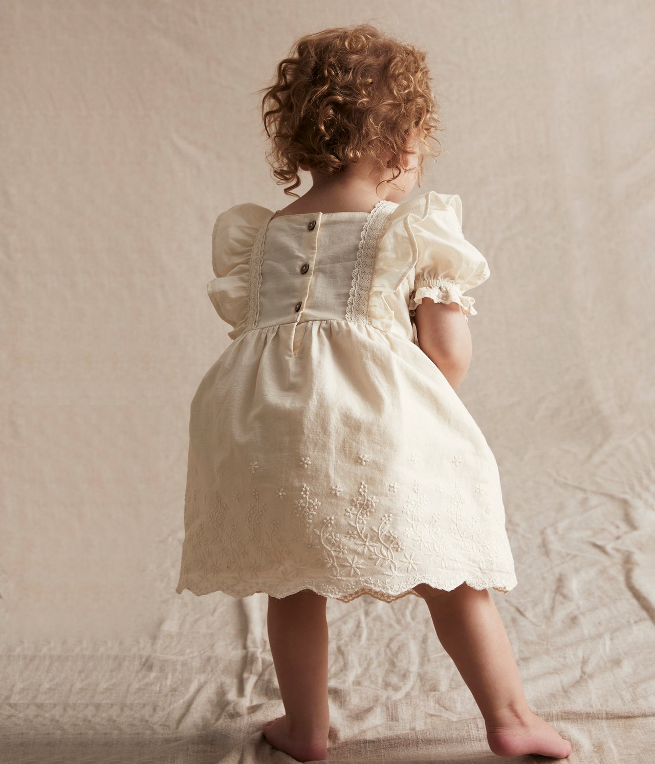 Back view of a baby wearing an off-white embroidered cotton dress with puff sleeves, lace details, and back buttons.