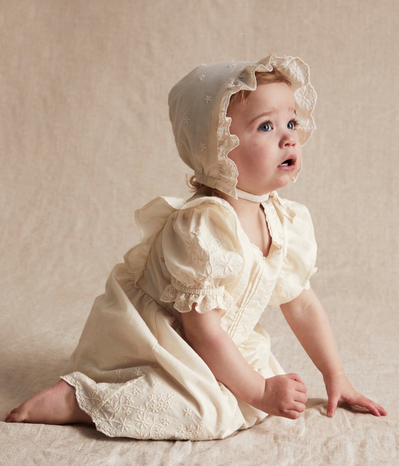 Front view of a baby in an offwhite embroidered cotton dress, puff shorts, and matching bonnet.