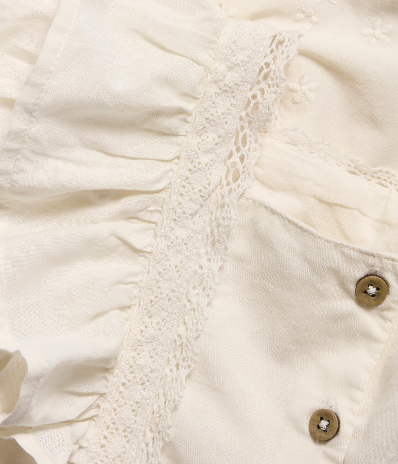 Close-up of an off-white woven cotton baby dress with lace trim, ruffles, embroidery, and two brown buttons.