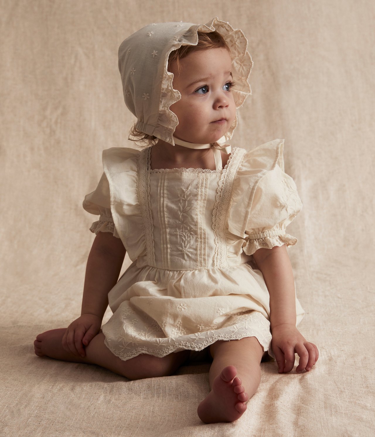 Front view of a baby wearing an off-white embroidered cotton dress with puff sleeves and a matching bonnet.