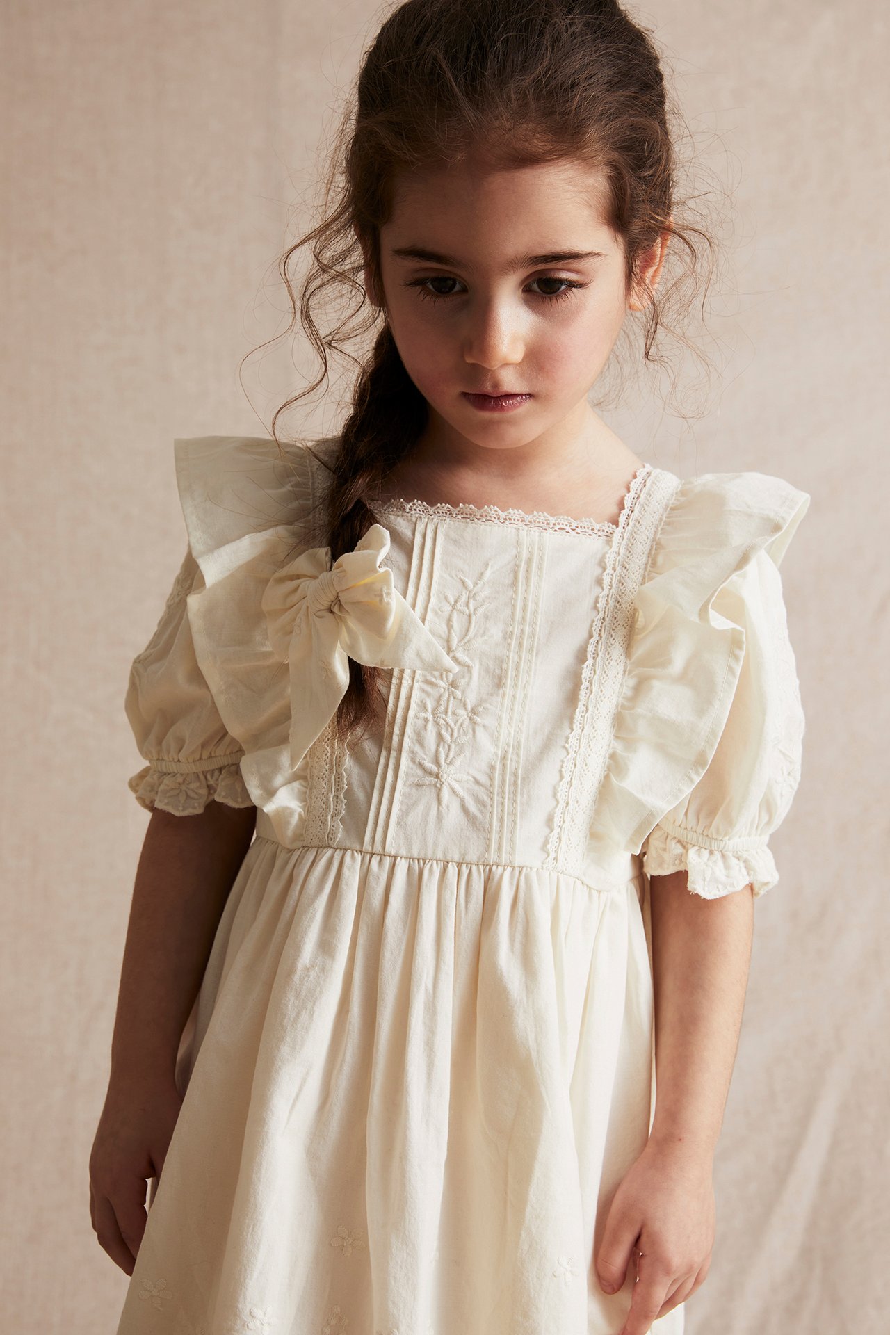 Front view: Kid in an offwhite woven cotton dress with lace, embroidery, frills, puff sleeves, and a bow.