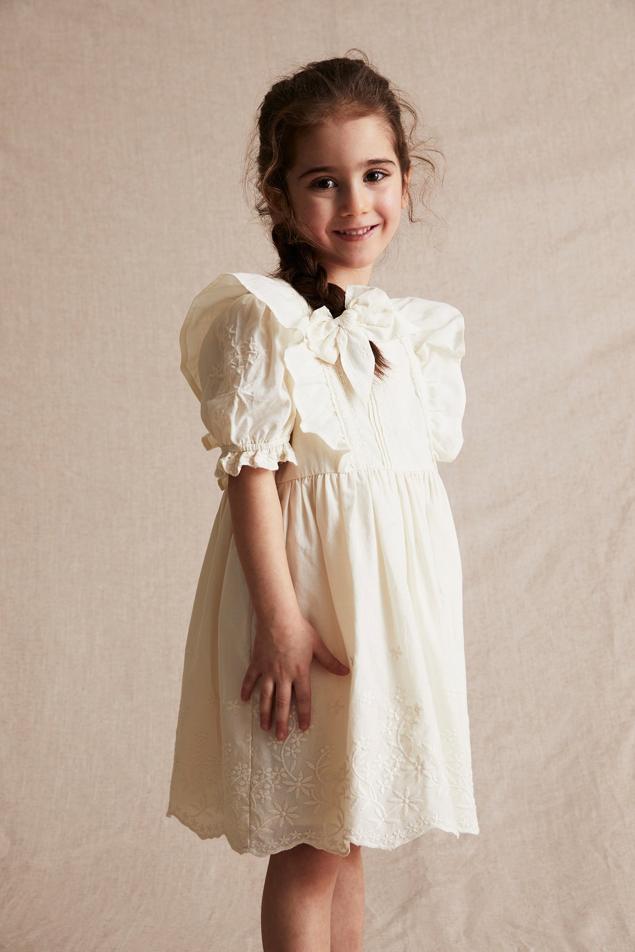 Front view of a smiling kid wearing an offwhite embroidered cotton dress with puff sleeves and shoulder frills.