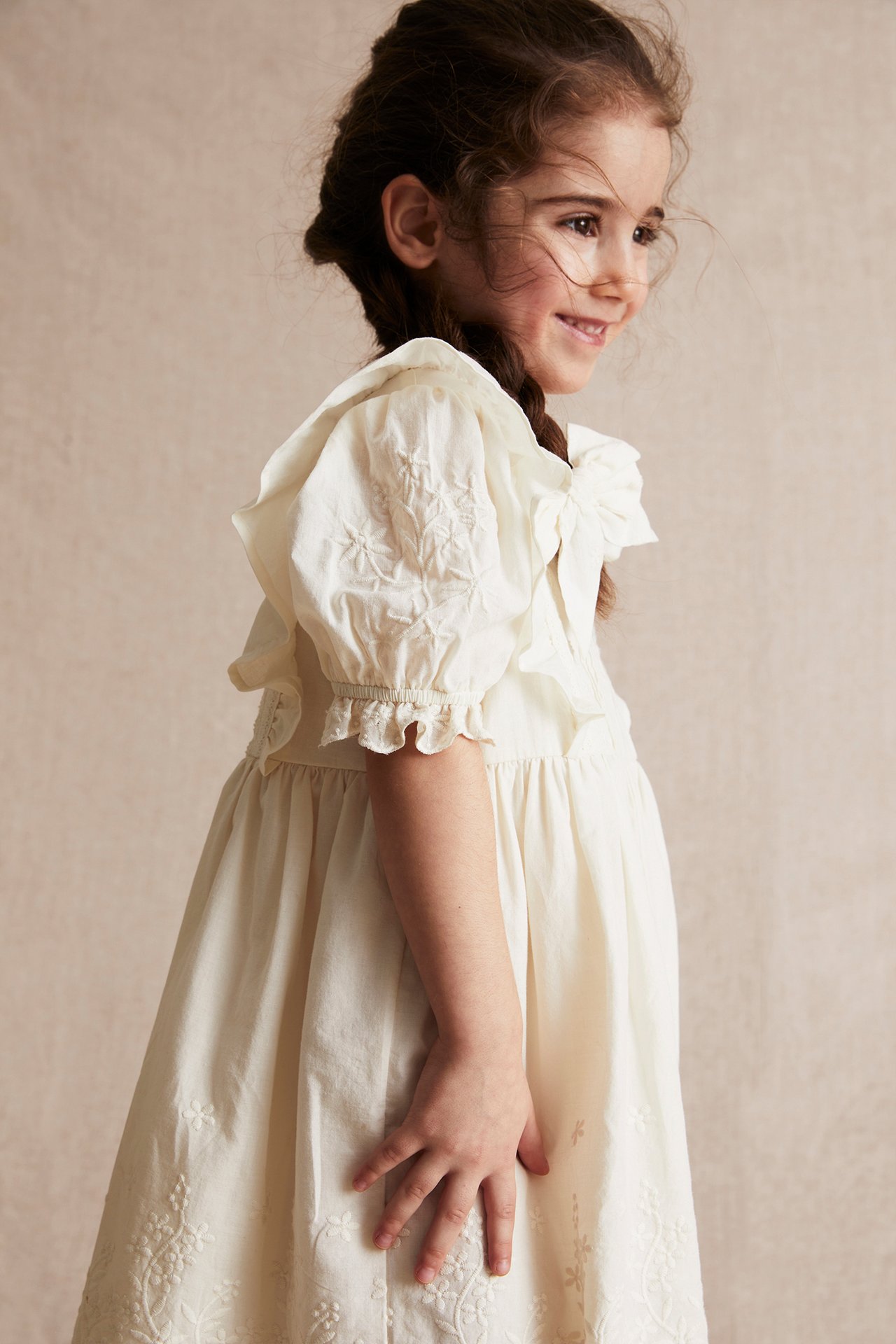 Side view of a kid wearing an offwhite woven cotton dress with puff sleeves, shoulder frills, and embroidery.