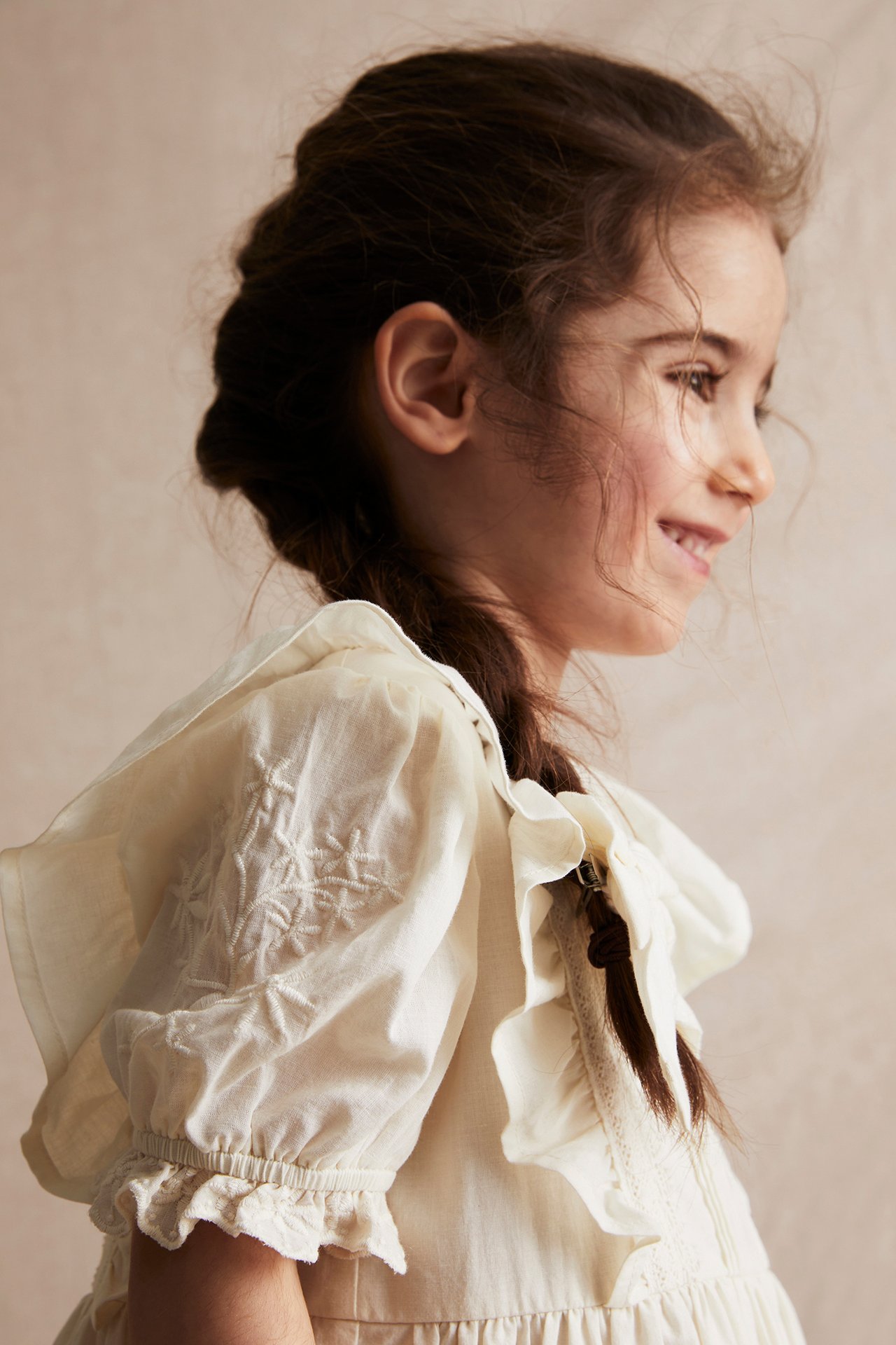 Close-up side view of a kid wearing an off-white cotton dress with embroidered puff sleeves and shoulder ruffles.