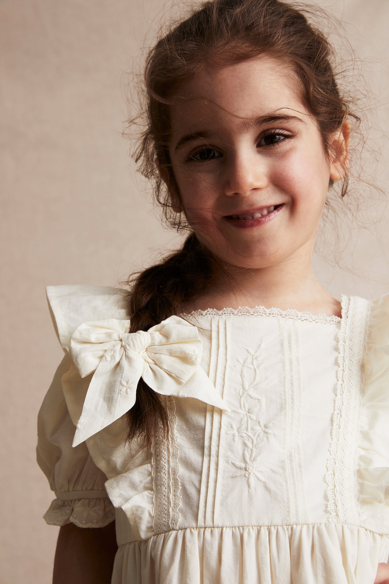Close-up front view: smiling kid in an offwhite woven cotton dress with lace, embroidery, and a bow.