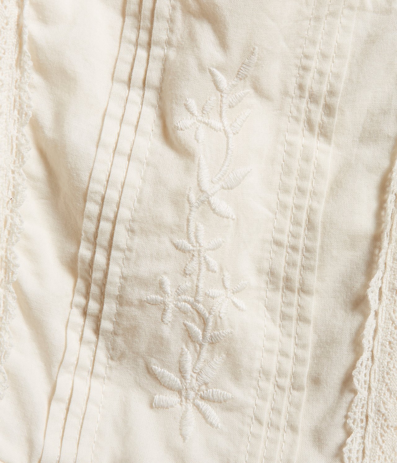 Close-up of an offwhite kid's woven cotton dress with pintucks, floral embroidery, and lace trim.