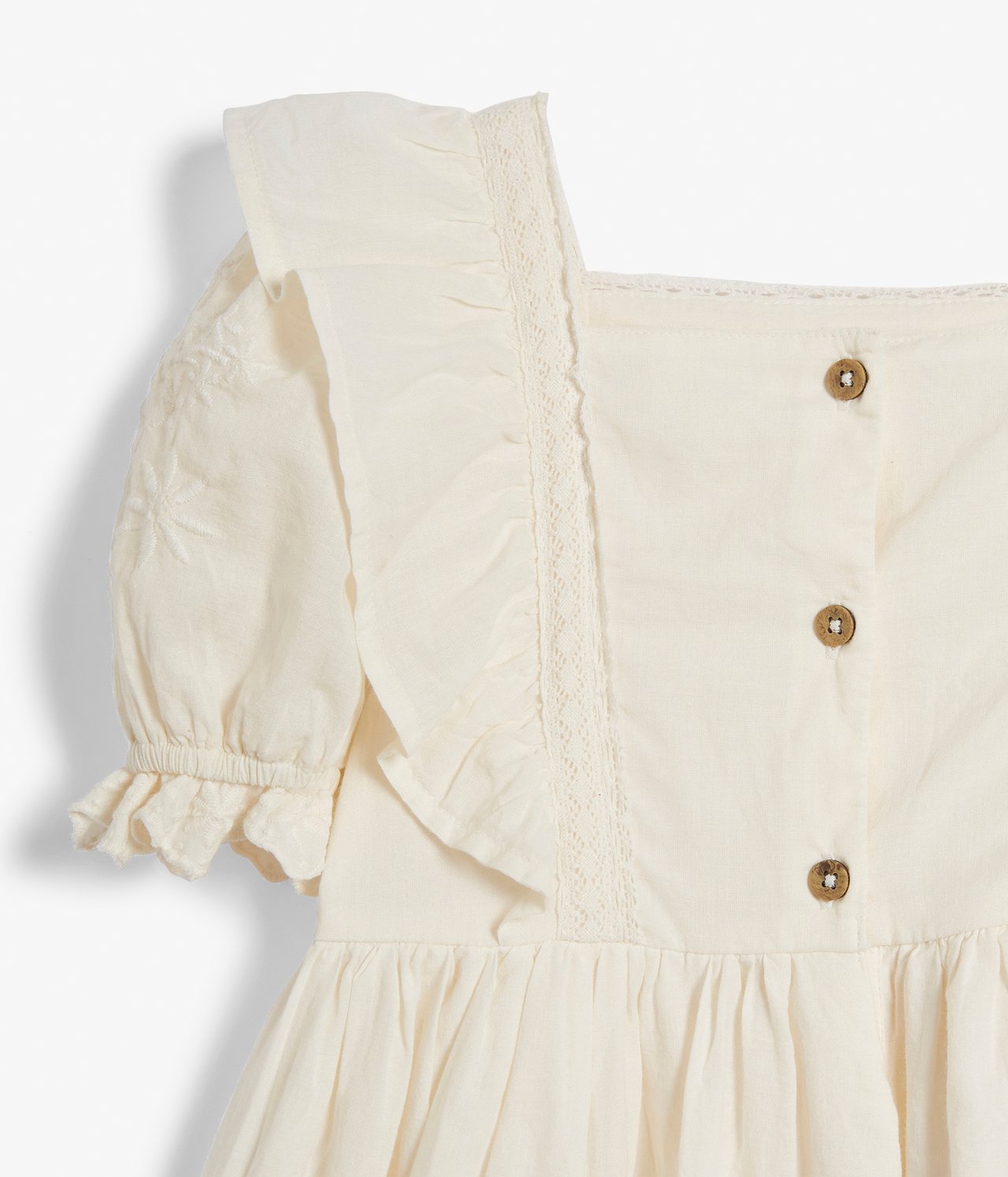 Close-up front view of an offwhite woven cotton kid's dress with lace, embroidery, frills, puff sleeves, and front buttons.