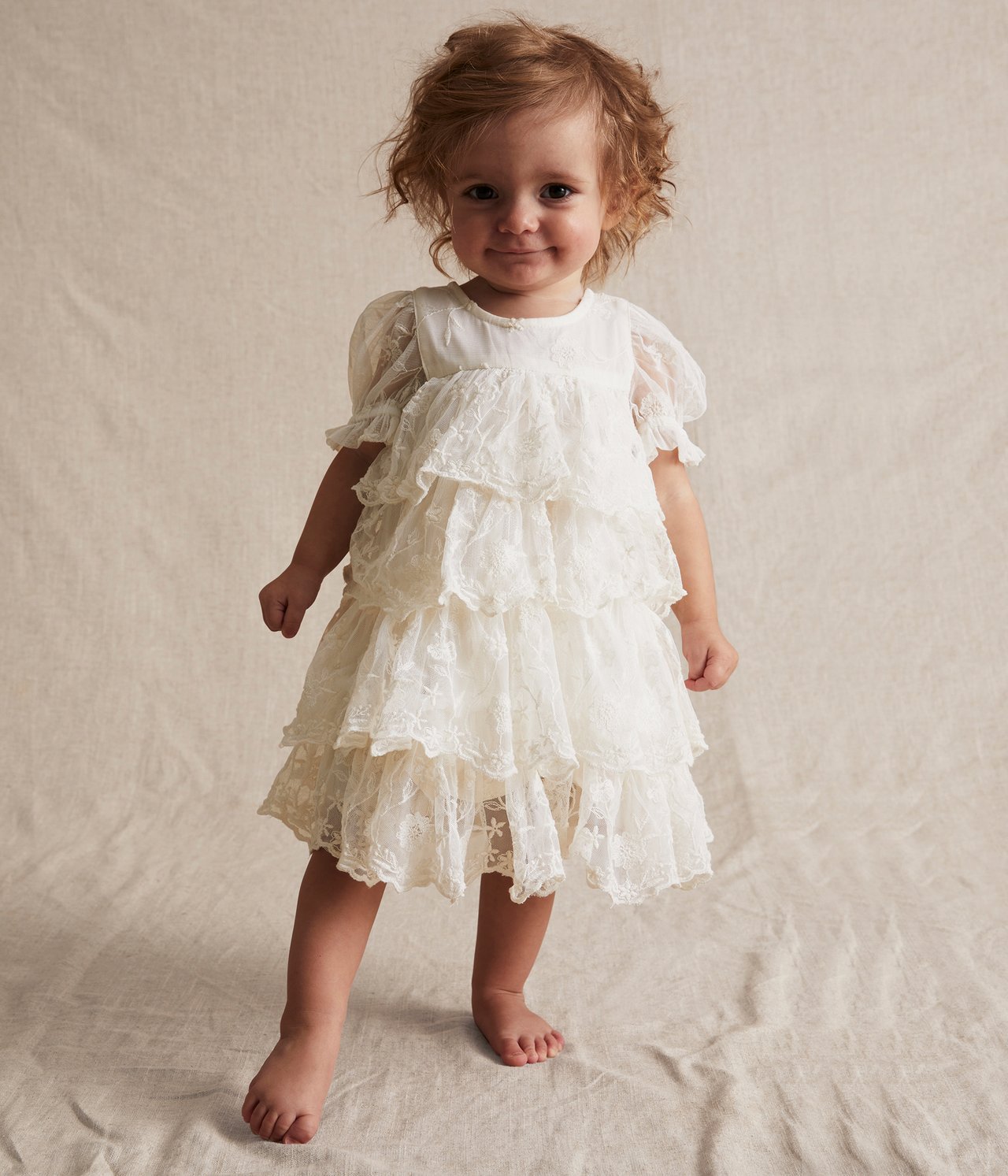Vorderansicht: Baby in einem cremeweißen, bestickten Mesh-Kleid mit Stufenrüschen und Puffärmeln.
