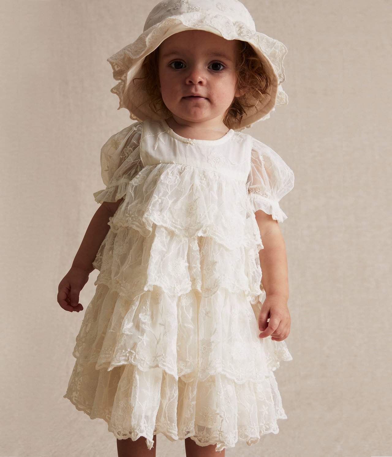 Vorderansicht eines Babys in einem cremefarbenen, bestickten Mesh-Kleid mit Rüschen und passendem Hut.