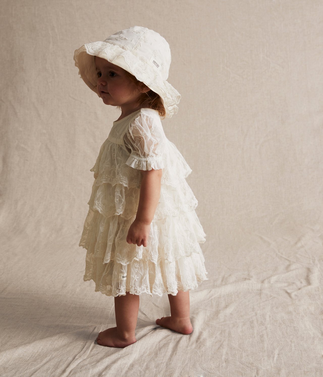 Seitenansicht eines Babys in einem cremeweißen, bestickten Mesh-Kleid mit Rüschen und passendem Hut.