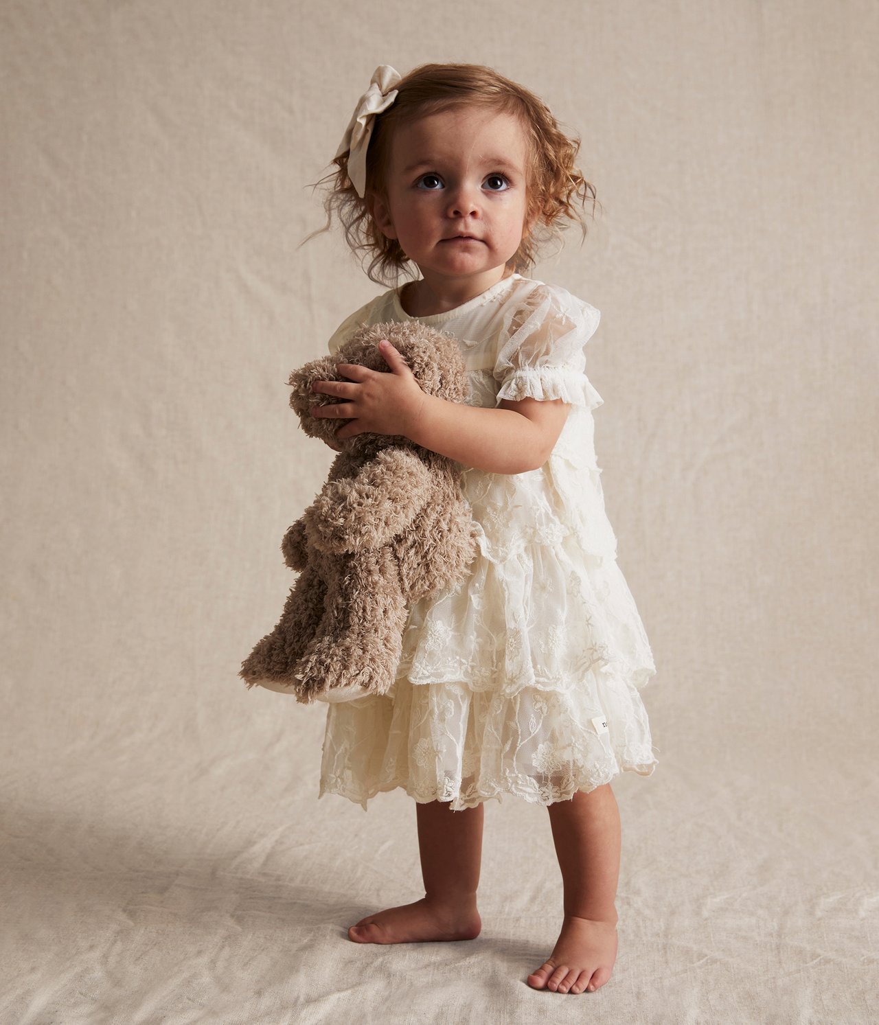 Vorderansicht eines Baby-Mädchens in einem cremefarbenen bestickten Mesh-Kleid mit Rüschen, hält einen braunen Teddybär.
