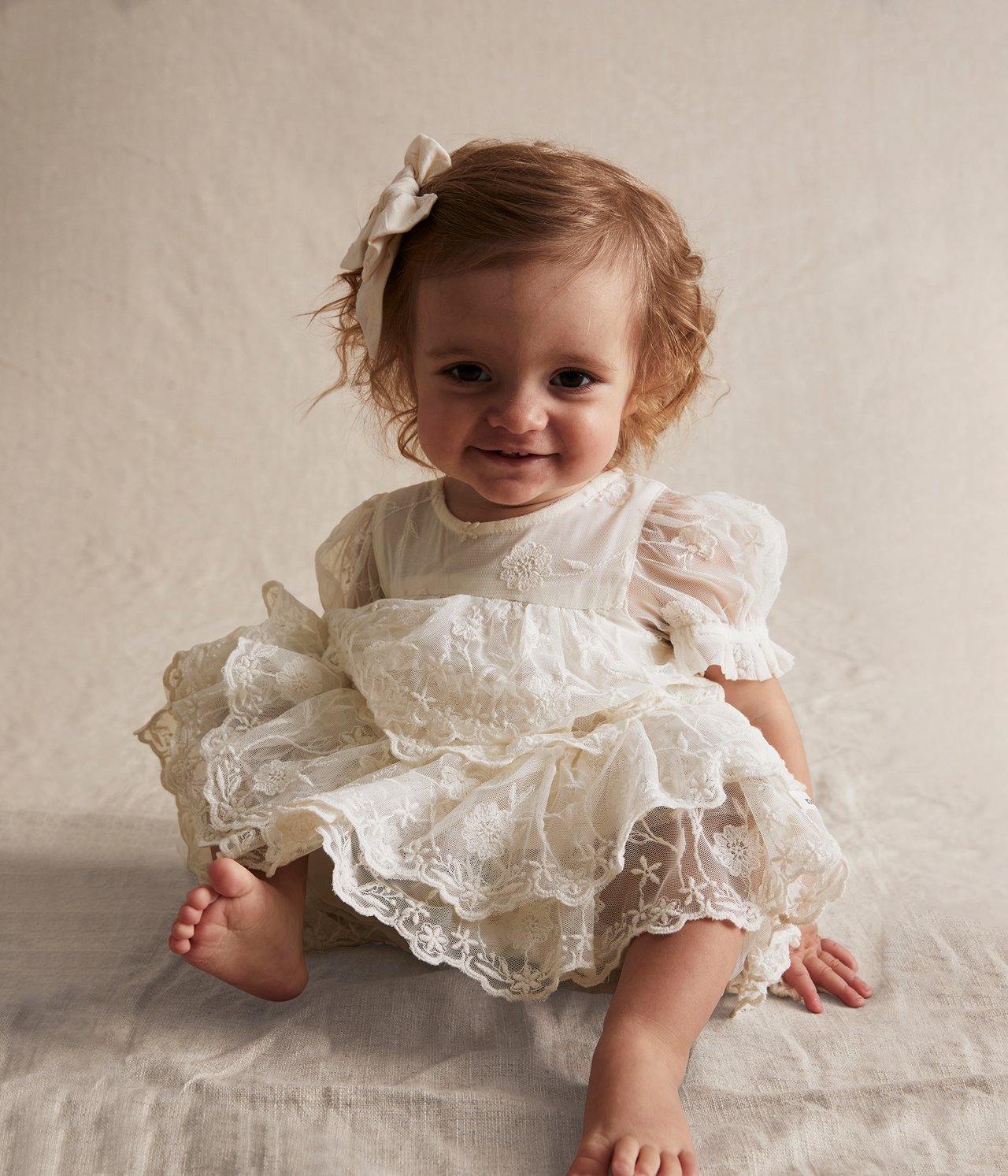 Vorderansicht eines lächelnden Babys in cremeweißem, besticktem Mesh-Kleid mit Puffärmeln und Haarschleife.