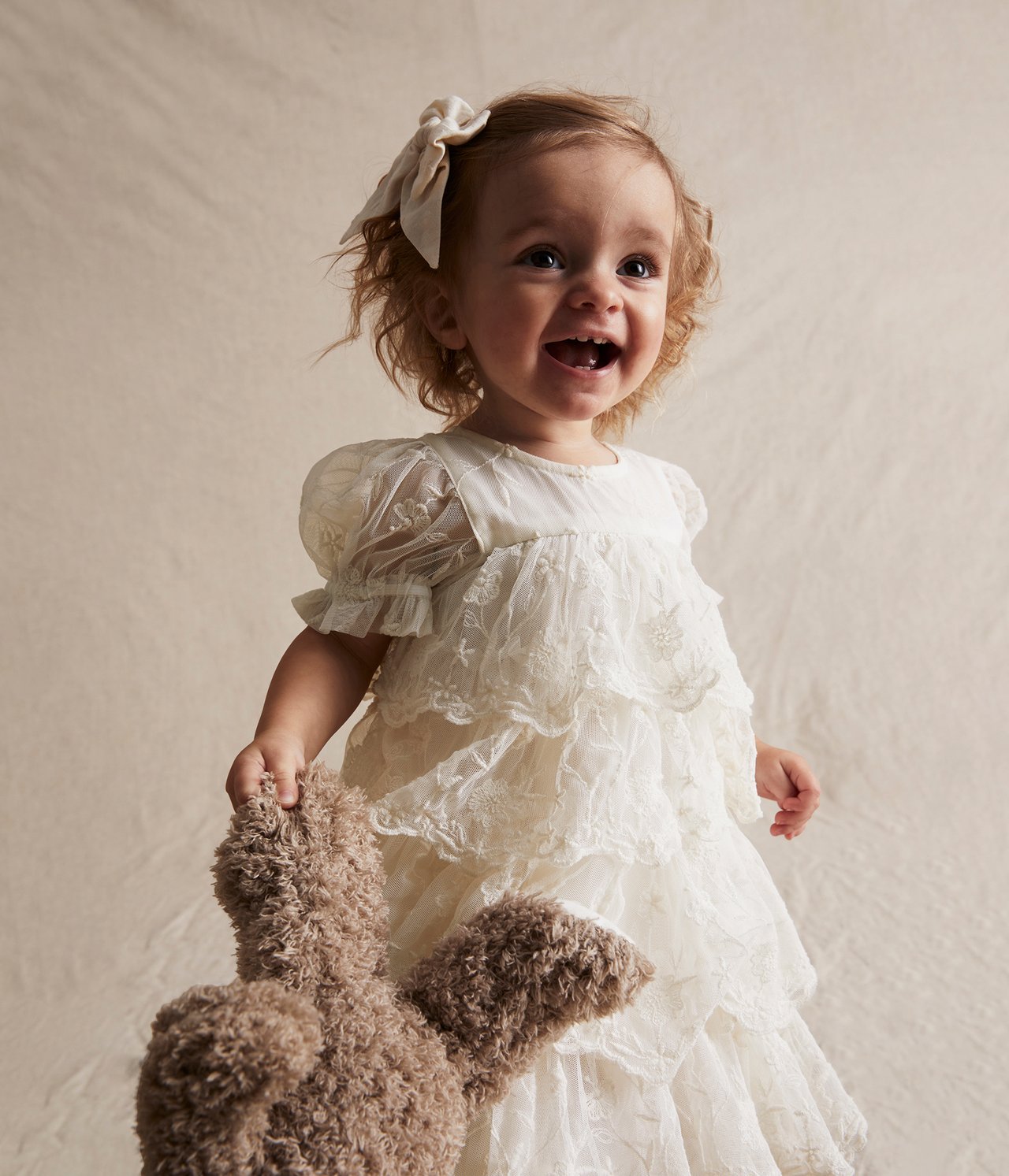 Vorderansicht: Baby in cremeweißem besticktem Netz-Kleid mit Puffärmeln, hält einen braunen Teddybär.