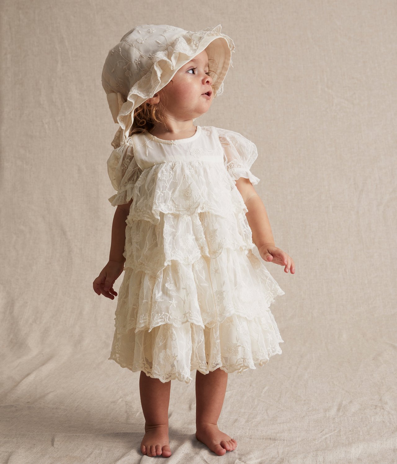 Vorderansicht: Baby in cremefarbenem besticktem Mesh-Kleid mit Stufenrüschen, Puffärmeln und passender Haube.