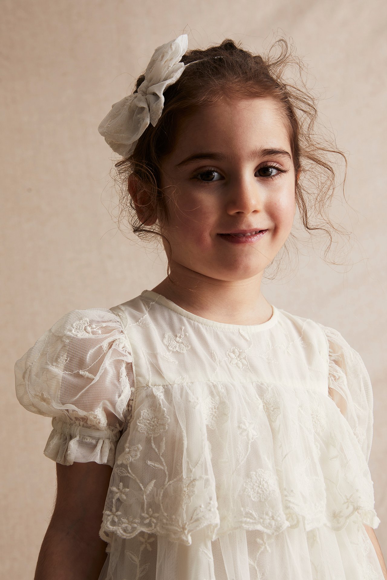 Close-up front view of a kid wearing an offwhite embroidered mesh dress with puff sleeves and a white bow.