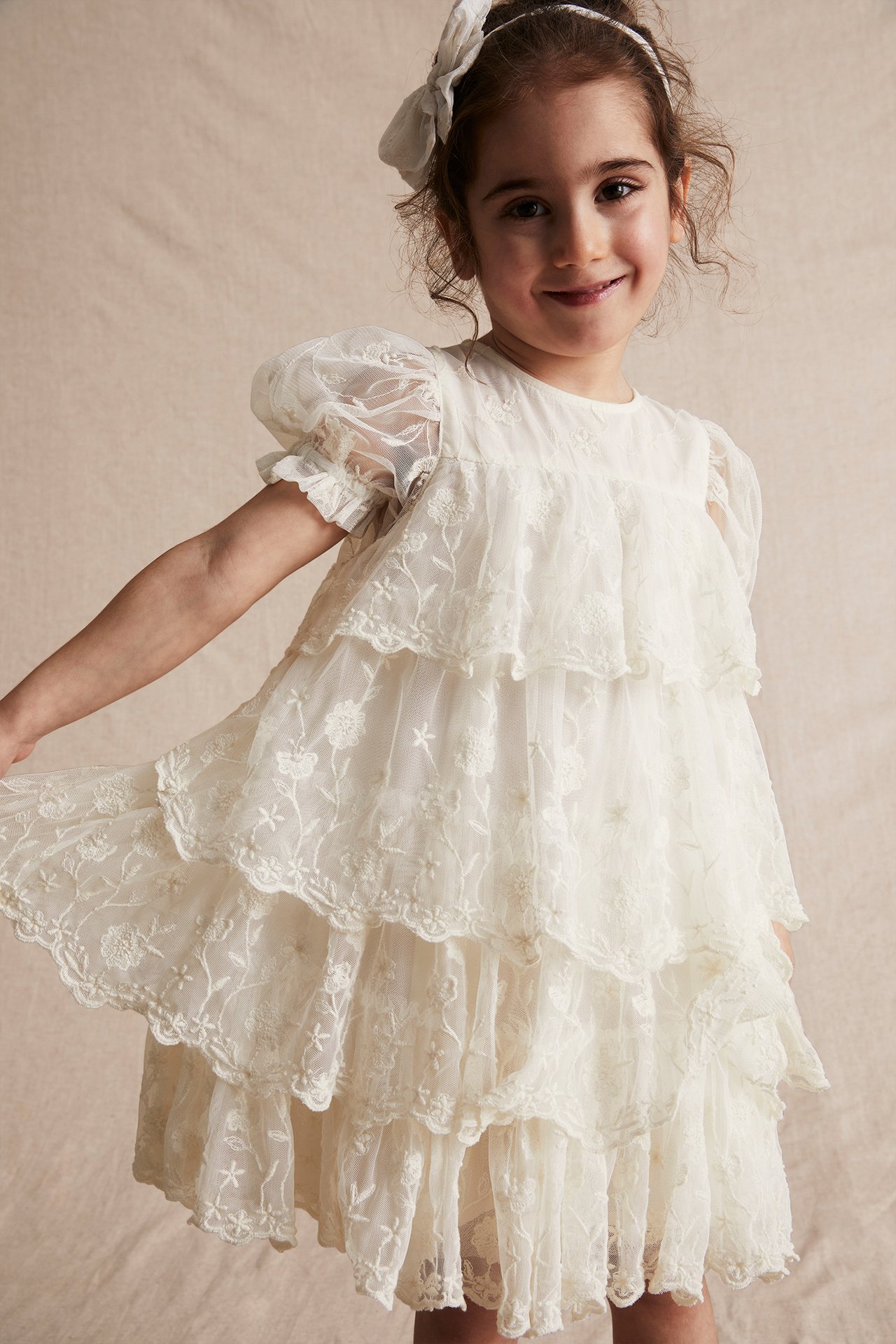 Front view of a kid in an offwhite embroidered mesh tiered dress with puff sleeves and a white bow.