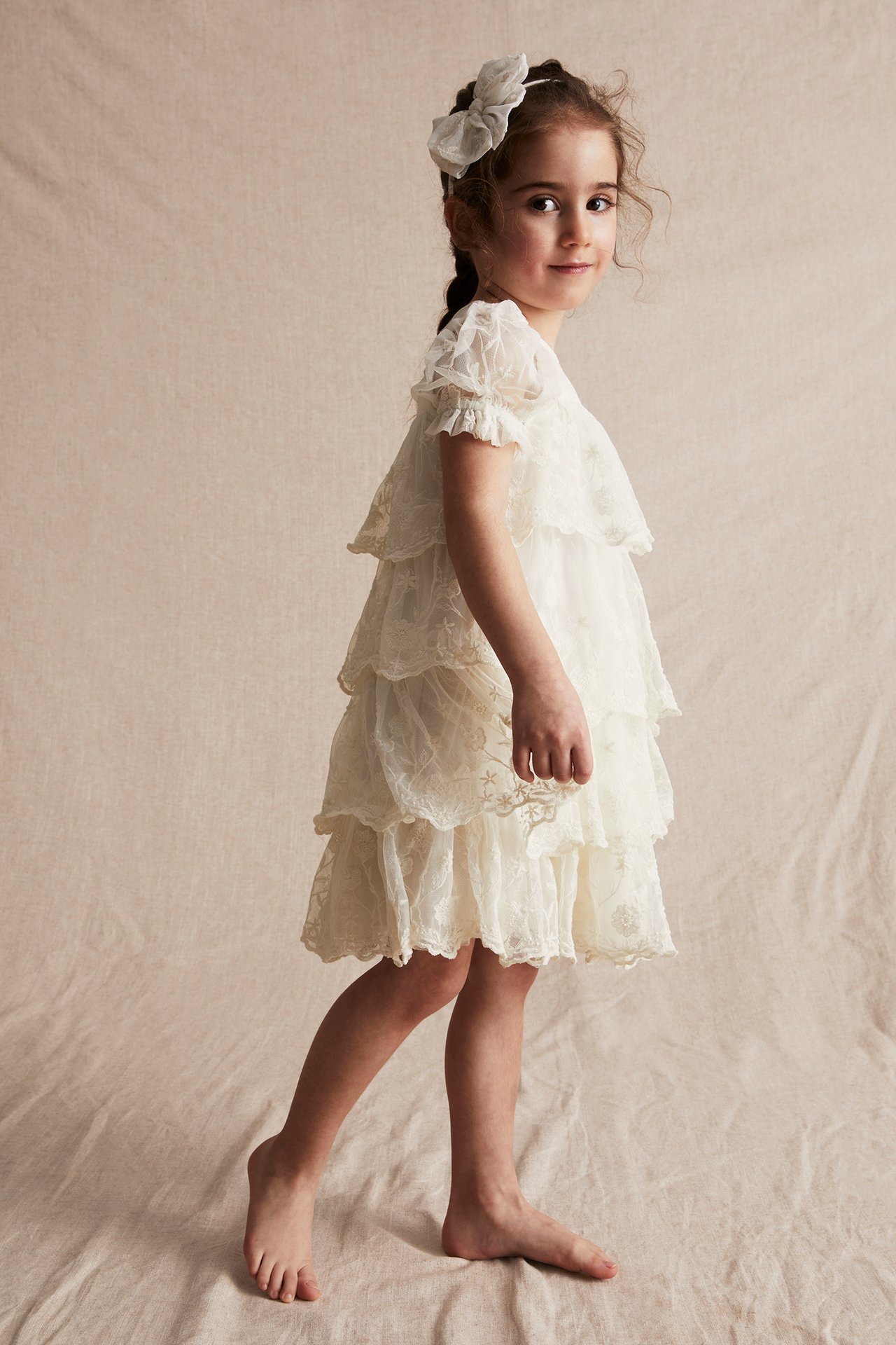 Side view of a kid wearing an offwhite embroidered mesh tiered dress with puff sleeves and a matching bow headband.