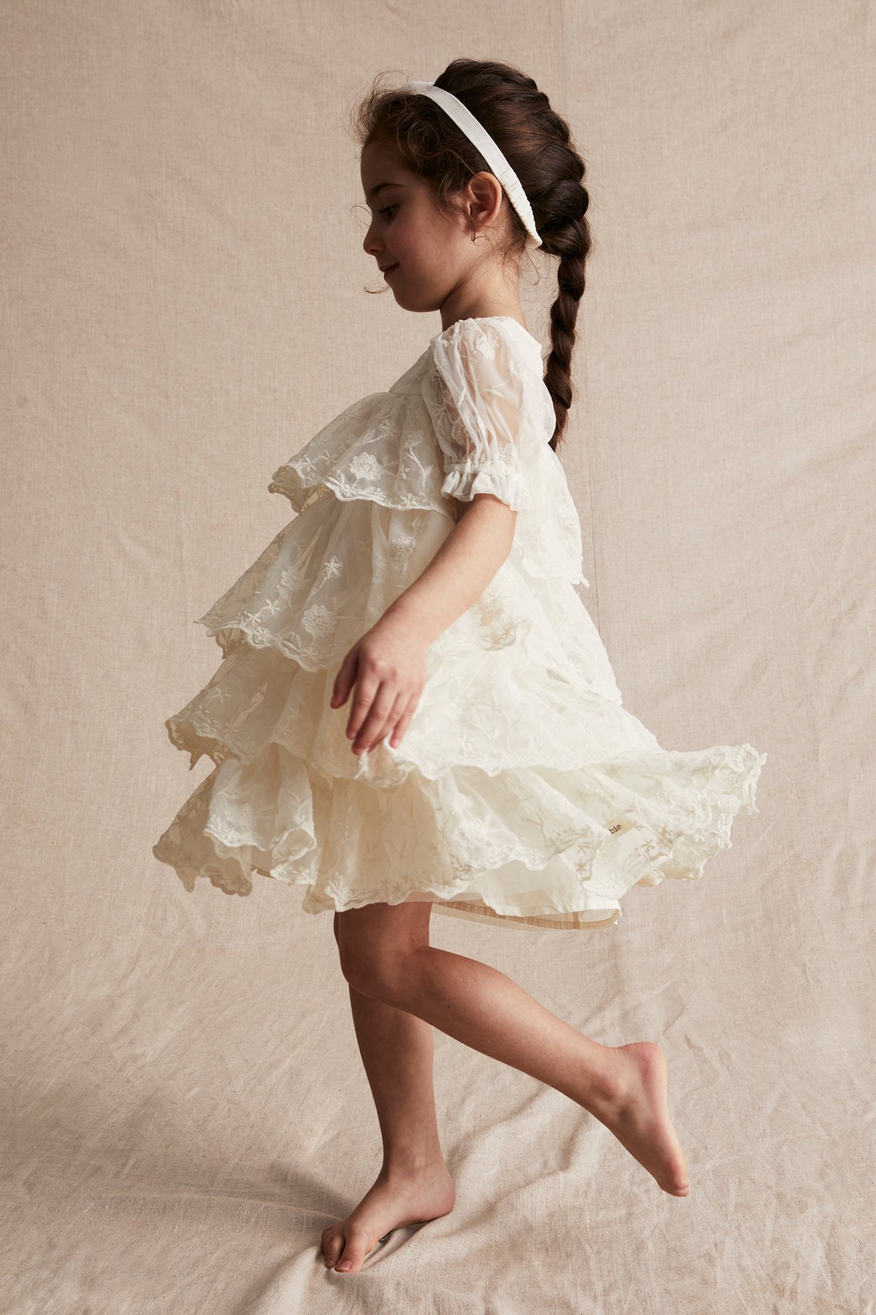Side view of a kid in an offwhite embroidered mesh dress with tiered ruffles, puff sleeves, and a white headband.