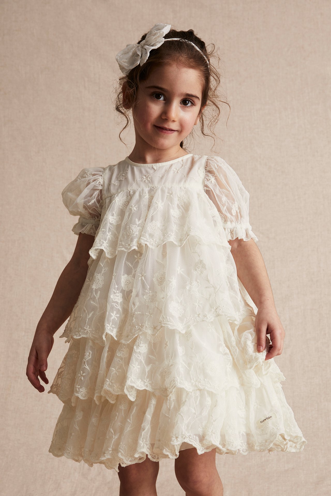Front view of a kid girl wearing an offwhite embroidered mesh tiered dress with puff sleeves and a white bow headband.