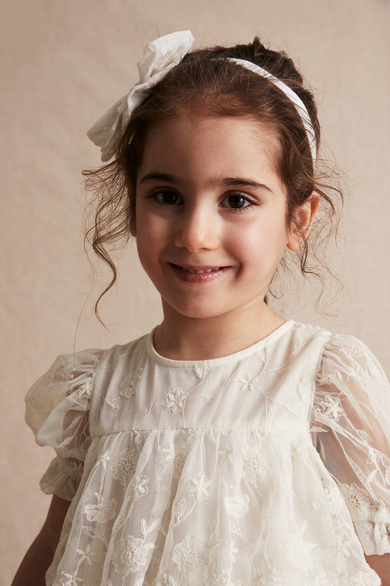 Close-up of a kid in an offwhite embroidered mesh dress with puff sleeves and a white bow headband.