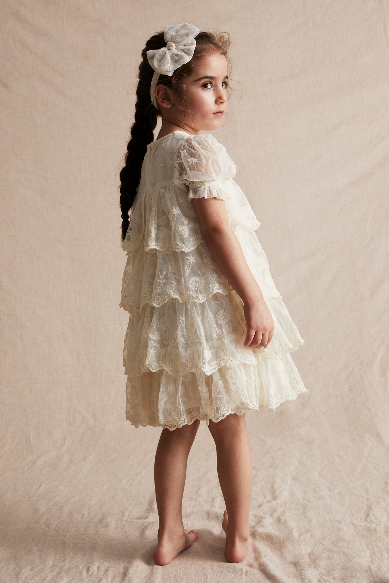 Side view of a kid wearing an offwhite embroidered mesh tiered dress with puff sleeves and a matching bow headband.
