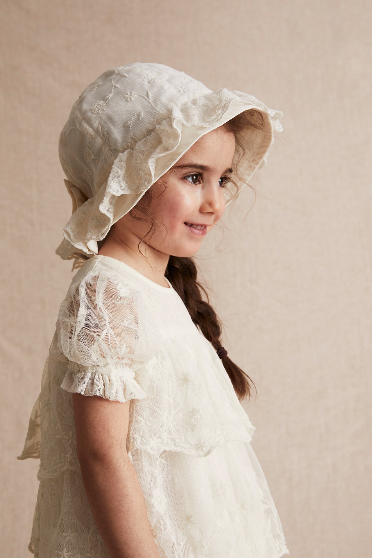 Side view of a kid wearing an offwhite embroidered mesh dress with puff sleeves and a matching embroidered bonnet.