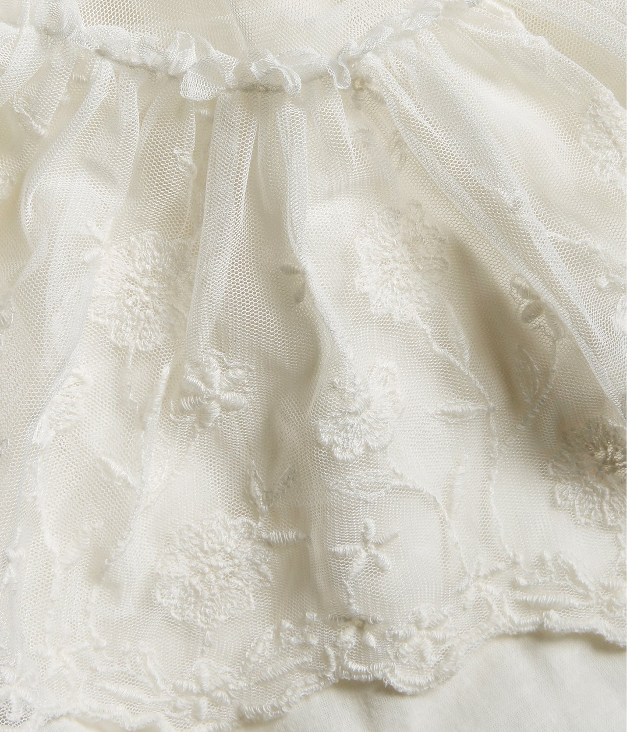 Close-up of an offwhite kid's embroidered mesh dress with ruffles and scalloped edges.