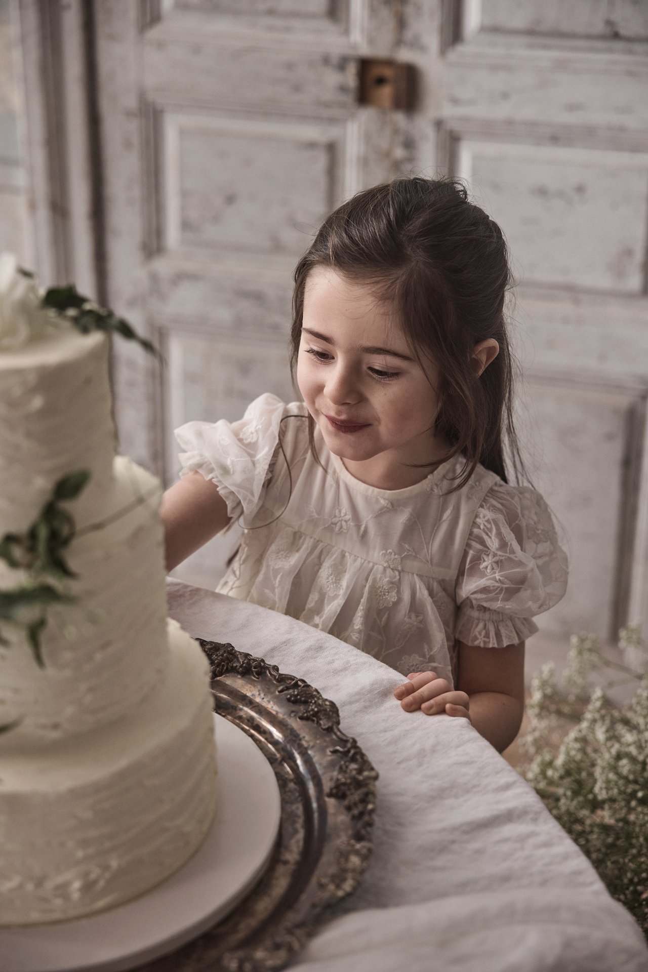 Front view of a kid in an offwhite embroidered mesh dress with puff sleeves, looking at a cake.